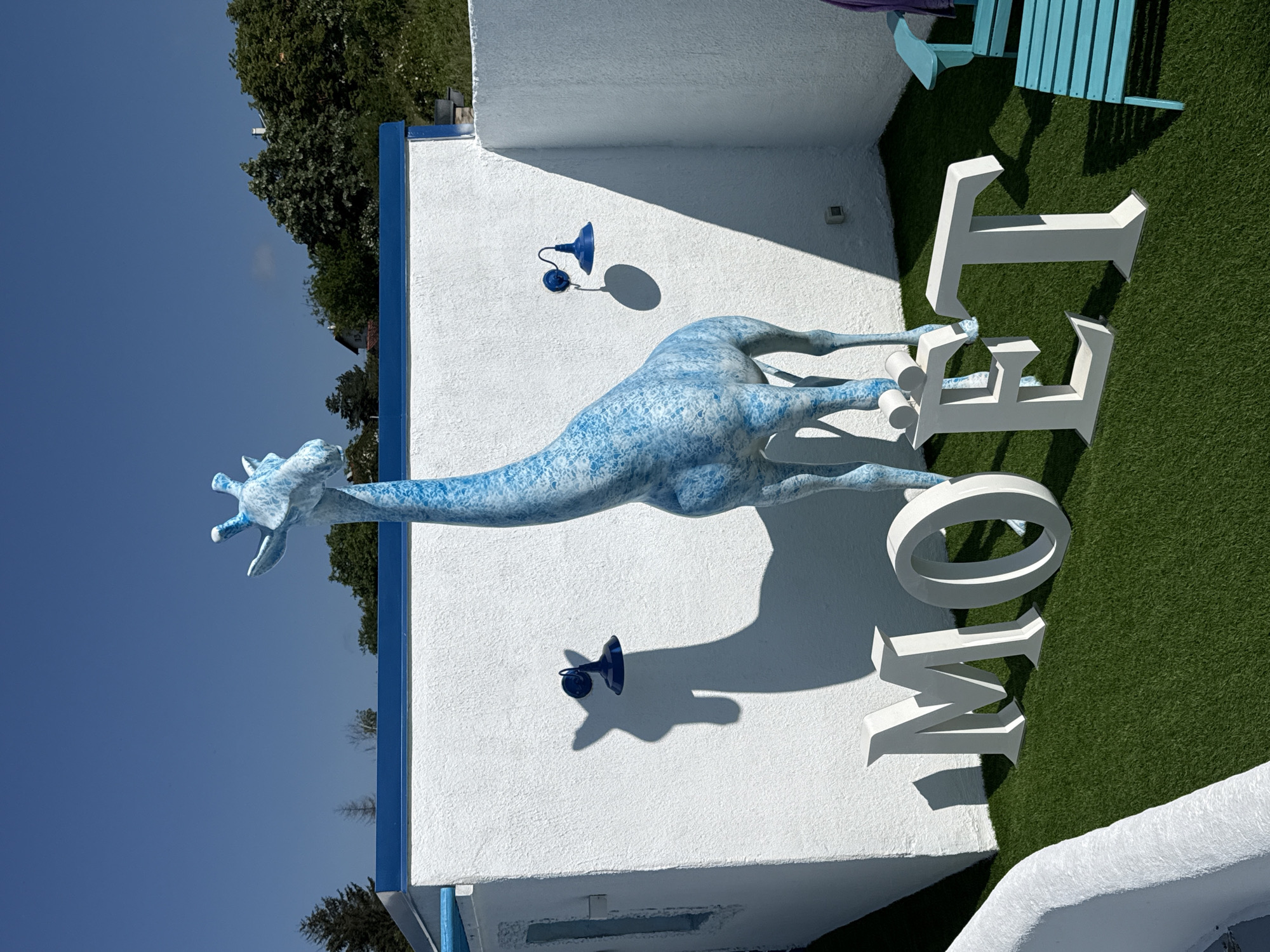 The image depicts an outdoor scene in Baliovtsi, Bulgaria, under a clear blue sky. A large, blue and white speckled giraffe statue stands prominently in the center of the frame. In front of the giraffe, large white letters spell out "MOET" on a patch of artificial grass. To the right of the statue, a blue adirondack chair with a purple towel draped over it is visible. Two dark blue wall-mounted lamps are fixed to a white textured wall behind the giraffe, one casting a distinct shadow of the giraffe's head and neck. The environment appears to be a well-maintained outdoor area, possibly a garden or patio, with white stucco walls surrounding the space.