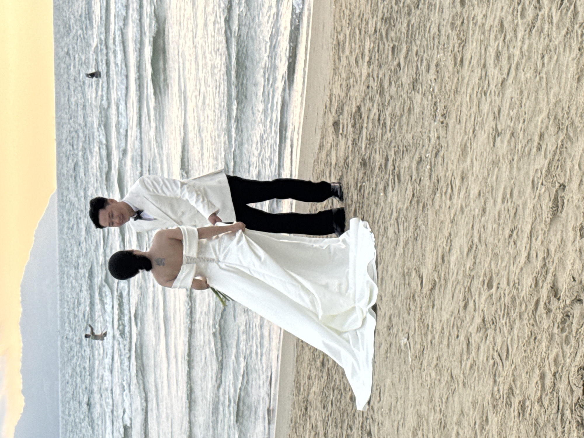 A newlywed couple is posing for a picture on a beach. The location appears to be Đà Nẵng, Vietnam, evident from the presence of the sea and land forms, with the Sơn Trà mountain range visible in the background. The groom, dressed in a white suit and black pants, is standing beside the bride. The bride is wearing a white wedding dress. The photograph is taken at the edge of the water, with the sea and the sand occupying a significant portion of the frame.