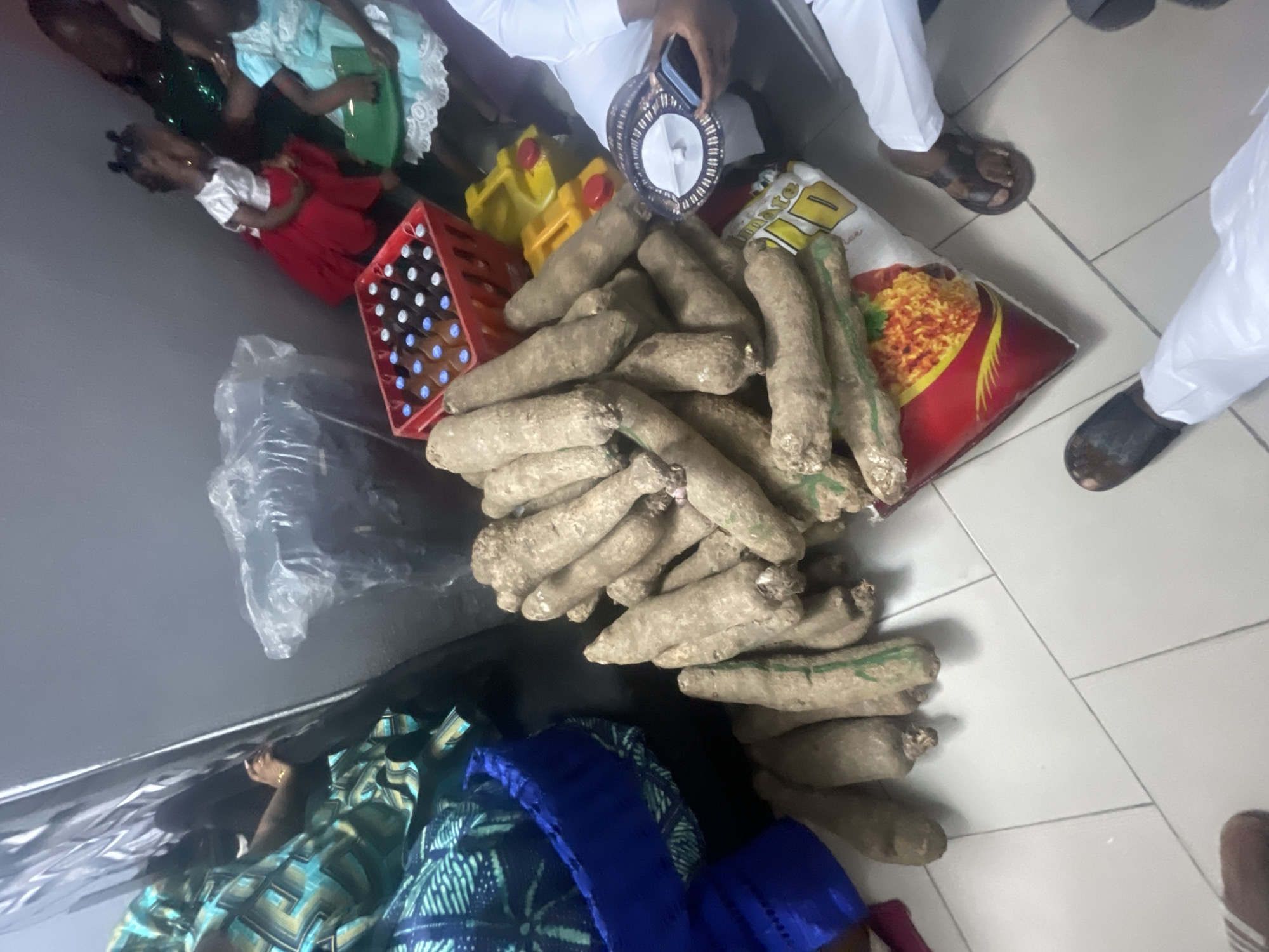 This indoor image from Lokoja, Nigeria, captures a moment centered around a distribution or gathering of food and beverages. In the foreground, a substantial pile of yams, a staple root vegetable, is arranged on a light-colored tiled floor.
Around the yams, several objects are visible: to the left, a red plastic crate filled with numerous brown bottles, likely containing drinks such as malt or soft drinks. Behind the yams, two yellow plastic jerrycans are placed. Further back, a large, colorful red and yellow sack of rice is prominent, with the partially visible brand name "DUMARTE" and an illustration of jollof rice on its front. A clear plastic bag lies on the floor to the left of the yams.
Several individuals are present, with only parts of their bodies visible. In the upper left, two young girls are seated; one wears a light blue dress with white lace trim, and the other a red dress with a white top. Adults are seen in white traditional Nigerian attire. One person is holding a mobile phone and a distinct circular patterned cap, typical of certain Nigerian cultures. Another adult's feet, wearing black sandals, are visible on the tiled floor to the right. Partial views of other individuals in patterned clothing are also seen on the lower left.
The setting is an indoor space characterized by light-colored, possibly white, tiled flooring and a dark, plain wall. The lighting appears artificial, suggesting it might be evening or an enclosed space. The overall scene conveys an organized arrangement of provisions, possibly for a communal event, a family celebration, or a charitable distribution within the Nigerian context. No specific emotions are discernible, but the scene appears calm and orderly.