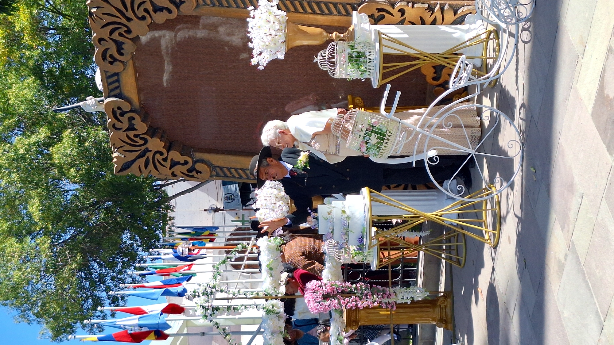 The image captures a scene in Cerro Colorado, Peru. An elderly couple dressed in formal attire stands beside a decorative white bicycle with gold accents, upon which is displayed a tiered cake and birdcages filled with flowers. The man wears a dark suit and hat, while the woman is in a light-colored outfit. A large, ornate frame is behind them. In the background, a row of colorful flags lines the street, with trees visible in the distance. The setting appears to be a public square or gathering place with paved walkways.