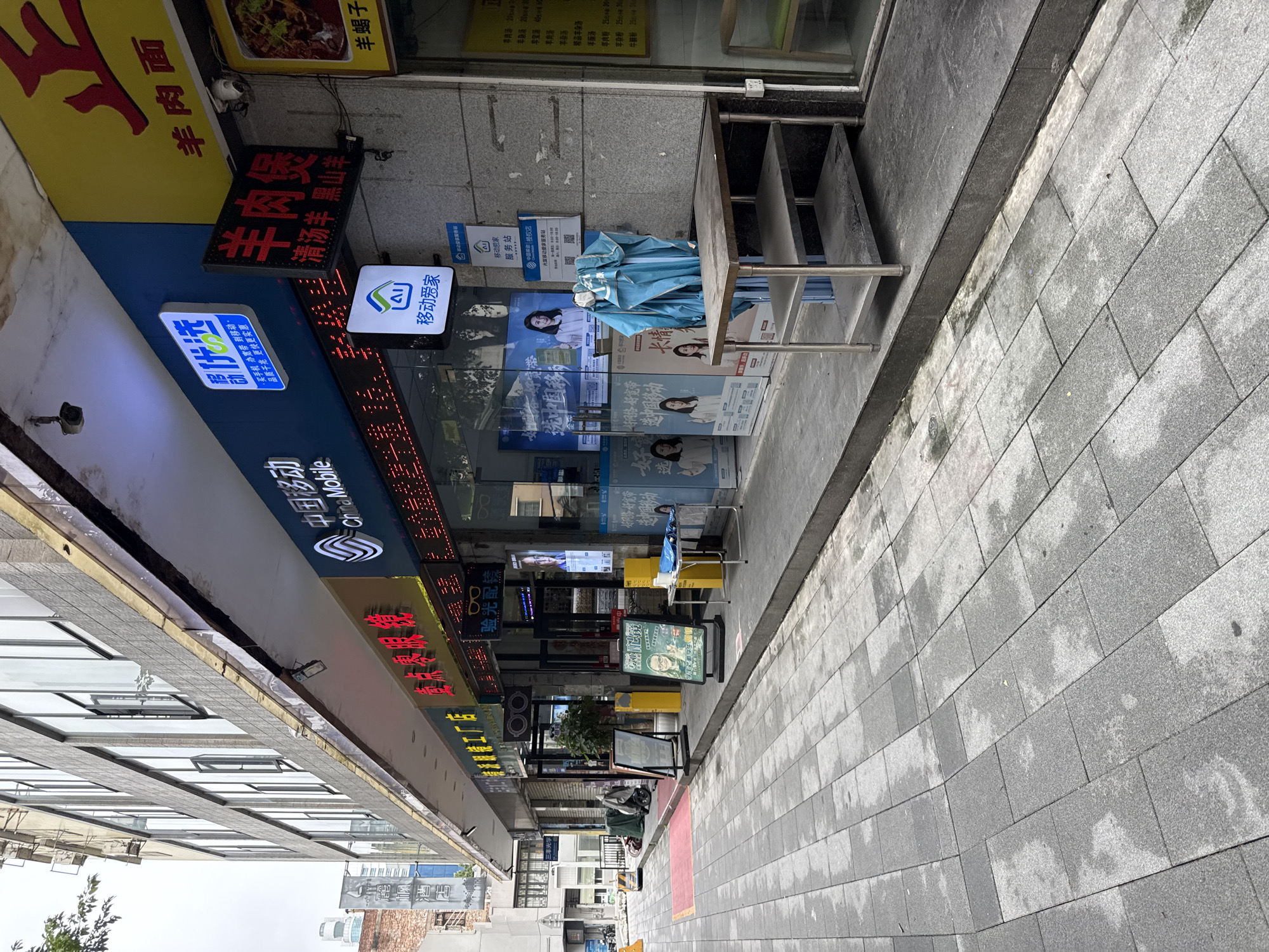 This image shows a street-level view of a commercial building facade in Liwan District, China. The building features multiple storefronts with illuminated signage. To the left, a building with a textured gray facade has large windows. The sidewalk in front of the shops is paved with gray square tiles, some appearing wet.
The storefronts display various signs. One sign, a yellow banner, has red Chinese characters at the top and "羊肉面" (lamb noodle) below. To its left, a blue sign with white Chinese characters reading "移动优选" (Mobile Preferred) is illuminated. Further left, a red neon sign reads "验光配镜" (Optometry and Prescription Glasses). Another blue sign with a white icon and Chinese characters "移动友家" (Mobile Youjia) is visible. Large posters featuring women are displayed in the windows of one shop.
In front of one of the storefronts, there is a metal table with three shelves, appearing dusty. A blue tarp or cloth is draped over the top shelf. A potted plant is situated near the entrance of the glasses shop. Two illuminated A-frame signs stand on the sidewalk. One sign shows a woman with glasses.
In the background, another building with a gray facade and numerous windows is visible. The sky is overcast. No people are actively engaged in activities within the frame of the image, though posters suggest customer interactions. The scene appears to depict a typical streetscape with retail establishments.