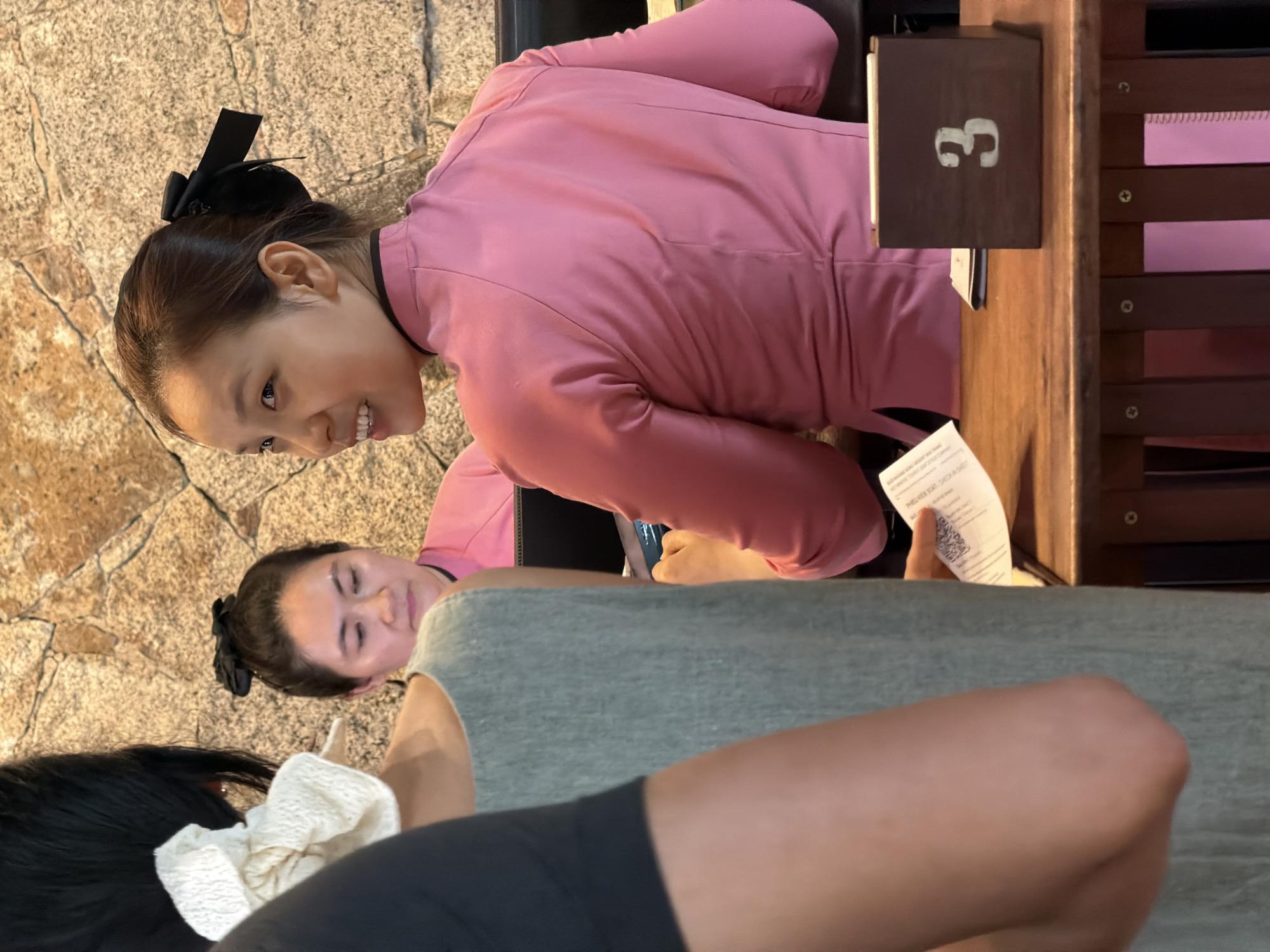 An over-the-shoulder shot captures a group of women interacting at a reception area, likely a hotel or restaurant in Nha Trang, Vietnam. The main subject, a woman in the foreground to the right, wears a pink, collared tunic with a black bow in her hair, and smiles as she looks away from the camera. Behind her, another woman in a similar pink tunic is partially visible, her head turned to the side. To the left, a person in a black shirt and a grey dress is seen from the back, facing the reception desk. A piece of paper with a QR code is being held by a person in a coral-colored top. A dark brown wooden box with the number "3" is placed on the wooden reception desk. The background features a textured stone wall.