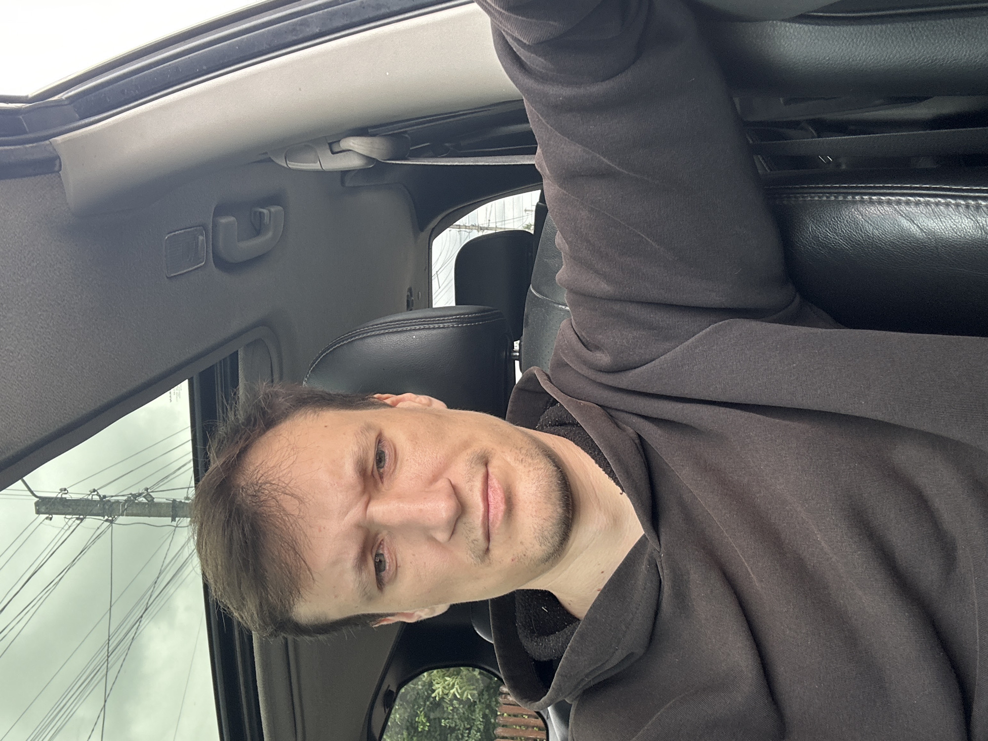 A selfie taken from inside a car shows a man sitting in the driver's seat. He is wearing a dark brown hoodie. The car's sunroof is open, revealing a cloudy sky with telephone poles and wires visible. The interior of the car, including the black leather seats and part of the dashboard, is also visible. The man has short brown hair, a serious expression, and stubble on his face. The image appears to have been taken outdoors in an urban or semi-urban setting. There are no other people visible in the image. The location is identified as Makiv, Ukraine, though there are no specific visual cues in the image to confirm this.