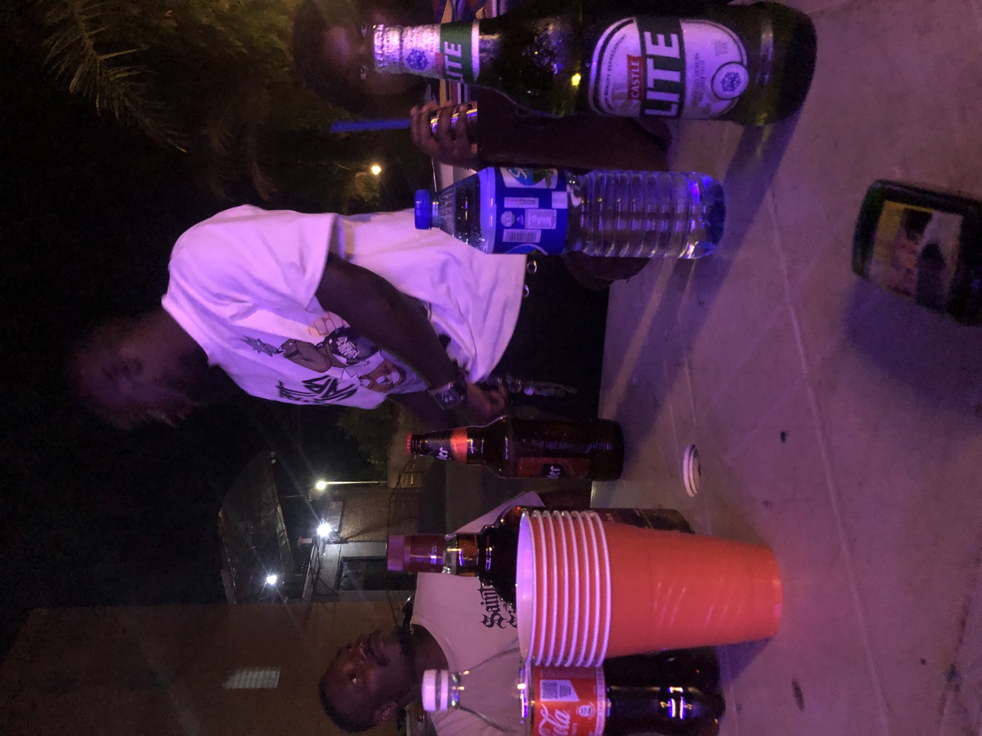 In an outdoor, nighttime setting, four individuals are gathered around a table with drinks. A person on the left, facing away from the camera, is looking upwards. Another person in the center, wearing a white t-shirt with a graphic print, is facing towards the left. To the right, a person is partially visible, holding a blue straw in a drink. A fourth person is likely present but not clearly depicted. On the table, there are several bottles of beverages, including a Castle Lite beer bottle and what appears to be a bottle of Coca-Cola. Red plastic cups are stacked on the table, and a bottle of water is also visible. The background shows a building structure and some foliage. The lighting suggests a social gathering, possibly at night.