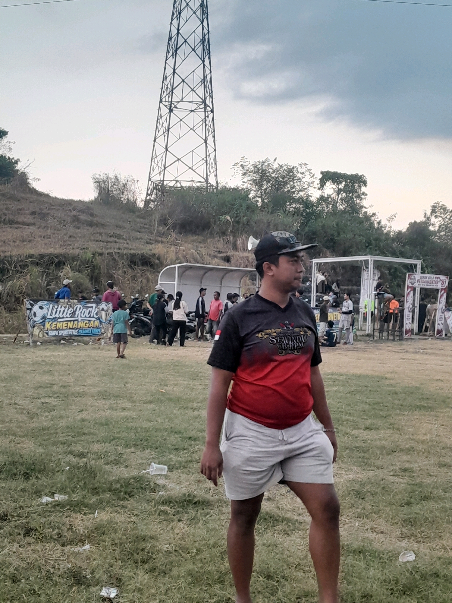 This image captures an outdoor scene with a man in the foreground standing on a grassy field. He is wearing a black and red t-shirt with a graphic design and light-colored shorts, and a black baseball cap. Behind him, a crowd of people are gathered near a banner that reads "Little Rock Kemenangan Tanpa Sportivitas Pasarnya Rampas." Several motorcycles are parked behind the banner, and a white covered structure, possibly a spectator stand, is visible in the background. Further in the distance, a tall metal tower dominates the skyline. The environment appears to be a field with dry grass and some scattered litter. The overall setting suggests an event or gathering taking place in an open area. The image was taken in Genteng, Indonesia.