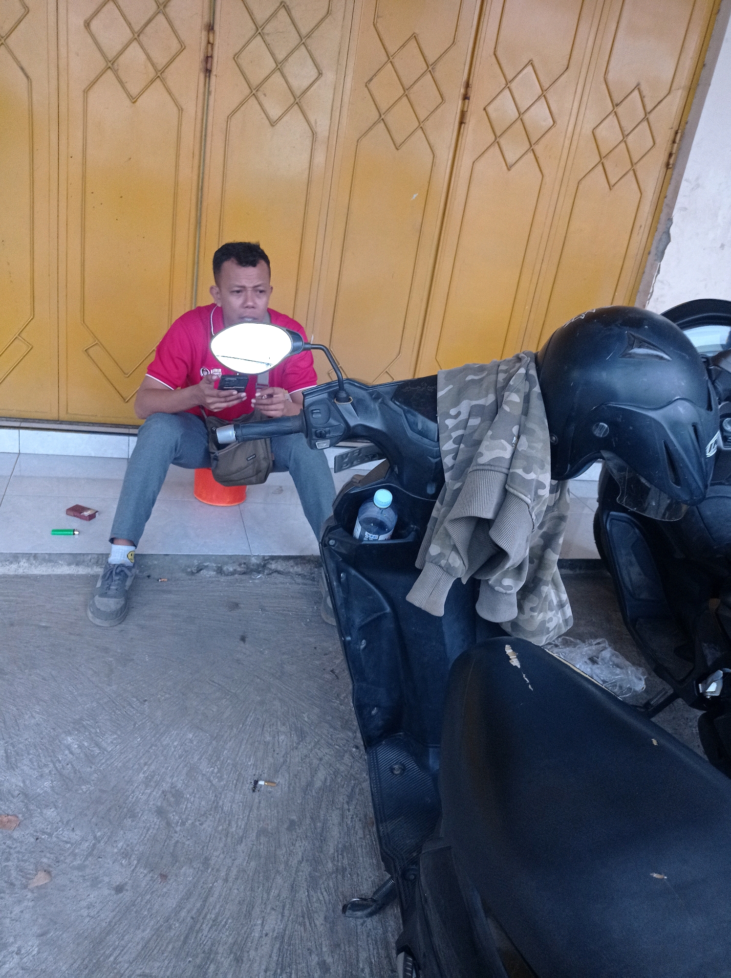 A man wearing a red shirt and grey pants sits on an orange bucket in front of a yellow, diamond-patterned door. He is holding a smartphone and a cigarette. A motorcycle with a black helmet and a camouflage jacket draped over it is parked in the foreground. A cigarette butt and a lighter are on the ground near the man's feet. The scene appears to be an outdoor area in Jetis, Indonesia.