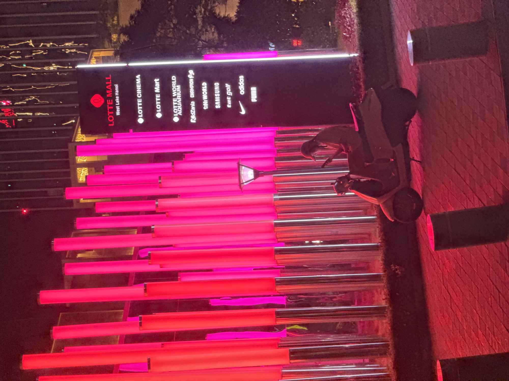 The image captures a nighttime scene outside a Lotte Mall in Hanoi, Vietnam. A tall, black sign lists various establishments within the mall, including Lotte Cinema, Lotte Mart, Lotte World Aquarium, KidZania, and several branded stores like Tini World, Samsung, Tat Golf, Adidas, and Puma. The sign is illuminated, as are rows of vibrant pink and red neon lights that line the walkway or building facade. In the foreground, a scooter is parked, its presence hinting at the mode of transportation common in the area. The overall atmosphere is modern and commercial, with the bright, colorful lighting creating a dynamic and inviting ambiance. The scene is likely set in an urban environment, given the presence of a large shopping mall and the style of architecture visible in the background. The time of day is clearly night, indicated by the artificial lighting and the darkness surrounding the illuminated areas.