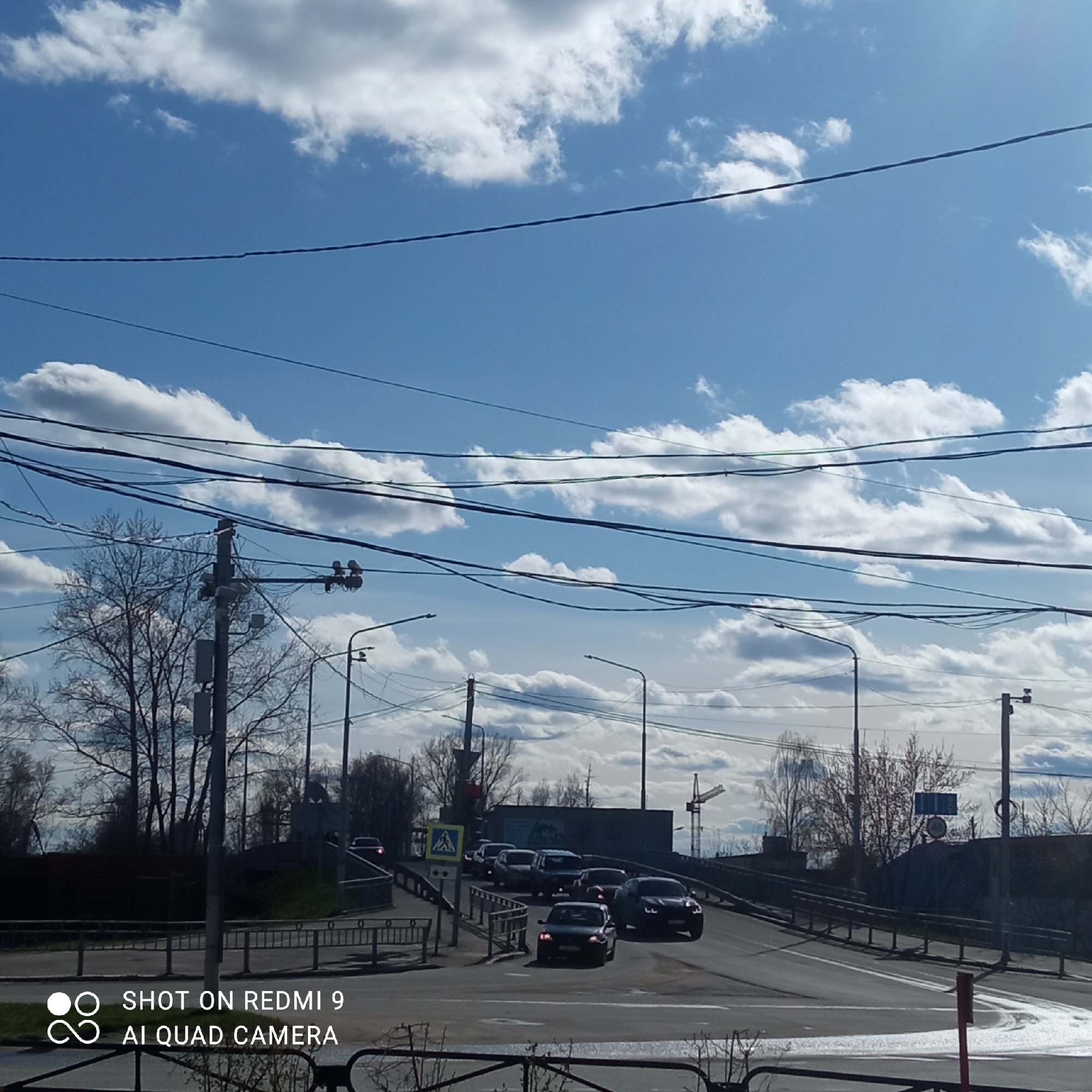 The image depicts a street scene in Chaikovsky, Russia. A bridge is visible with multiple cars traveling across it. A pedestrian crossing sign is present. Overhead, numerous power lines crisscross the sky, which is partly cloudy with patches of blue. There are bare trees and streetlights lining the road. In the background, a construction crane can be seen.