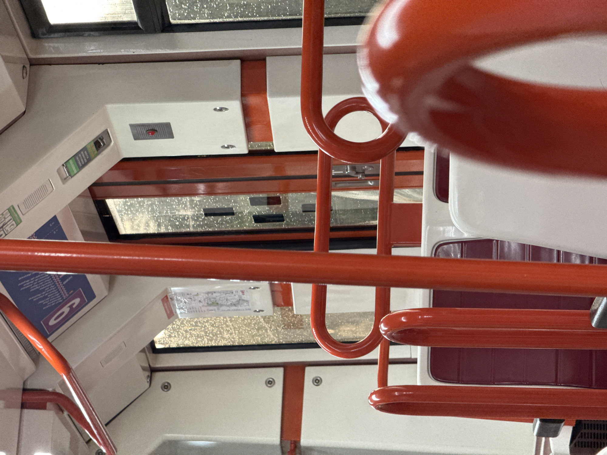 The image depicts the interior of a public transport vehicle, likely a train or metro, in Arganda del Rey, Spain. The scene is dominated by bright red handrails and seating, contrasting with the white interior walls and ceiling. Several red vertical poles and curved horizontal bars are visible, serving as handrails for passengers. Red seats with white trim are partially visible in the foreground and midground.
Through the windows, the outside environment is visible, appearing overcast with water droplets on the glass, suggesting it might be raining or the image was taken during a rainy day. The interior features various functional elements typical of public transportation, such as signage, buttons, and ventilation grilles. A blue and white sign with purple lettering, possibly a route map or informational display, is mounted on the upper wall. A red emergency button is visible on a white panel. The overall impression is one of a clean and functional interior of a modern public transport vehicle. No people are visible in the image.
