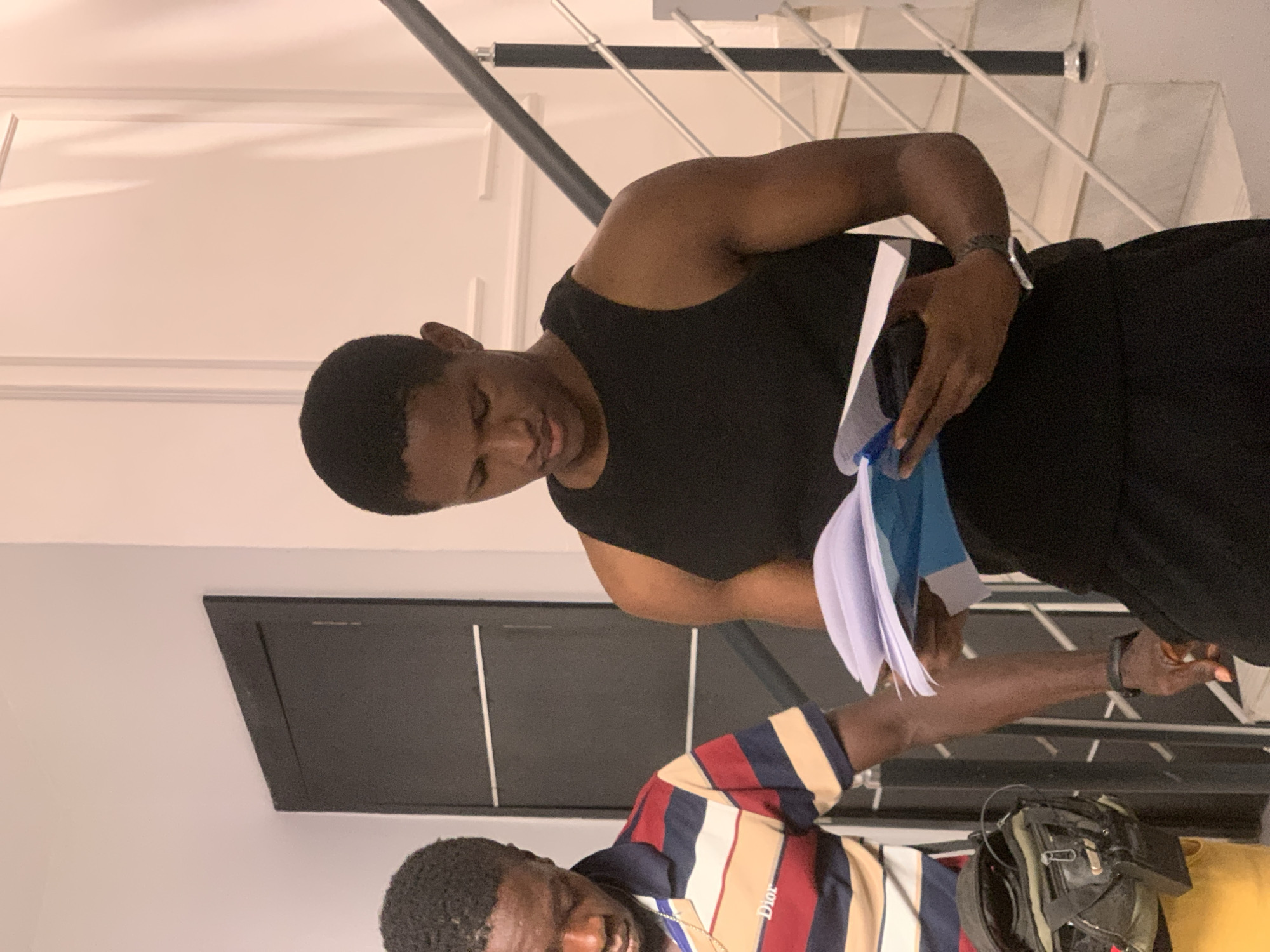 The image captures an indoor scene in Igboefon, Nigeria, featuring two young adult males. In the foreground, a male with short, dark hair, wearing a black sleeveless top and black pants, stands holding an open book or stack of documents with white and blue covers, looking downwards intently. A dark-colored smartwatch is present on his left wrist. Partially visible behind and to his left is a second male, also looking downwards, wearing a horizontally striped polo shirt in red, beige, dark blue, and light blue, with "Dior" text visible on the chest. He wears a gold-colored chain around his neck, and a dark green and black messenger bag is slung over his left shoulder or side. The background features a light-colored wall with decorative molding on the right, alongside a staircase with light-colored steps, a dark handrail, and white vertical balusters. On the left, a dark-colored door with horizontal metallic accents is visible against a light-colored wall.
