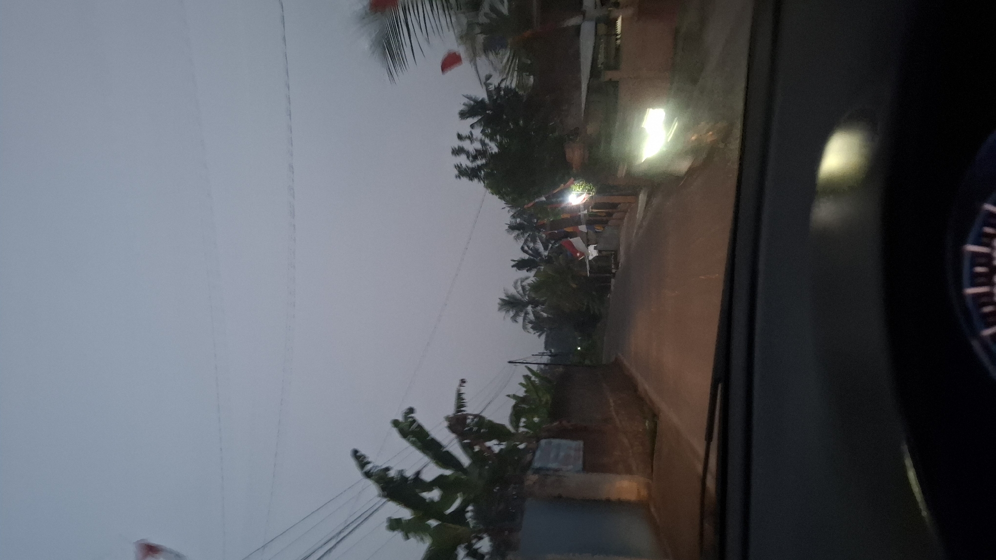 The image depicts a view from inside a vehicle looking out at a street in Cibinong Hilir, Indonesia, at dusk. The sky is a muted grey, suggesting either early morning or late evening. The road appears to be paved and is empty of vehicles or pedestrians. On either side of the road, there are houses with fences and lush vegetation, including banana trees and palm trees. Some houses have small flags displayed, possibly indicating a local celebration or holiday. A bright light from an oncoming vehicle or a street lamp illuminates the right side of the road. The foreground shows the dashboard of the car, with a portion of the steering wheel visible at the bottom. The overall impression is a quiet, suburban street scene in Indonesia as daylight fades.