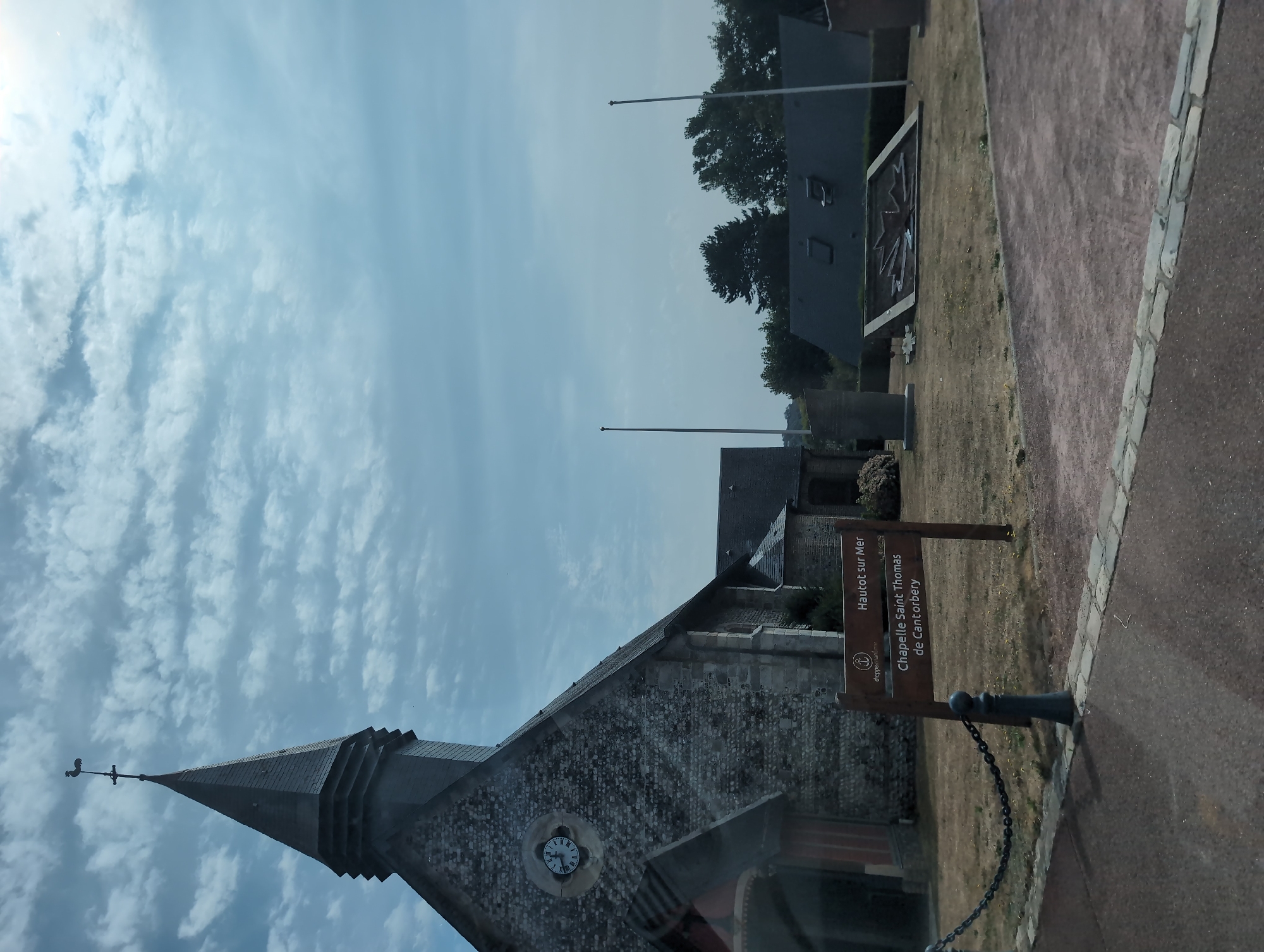 In Hautot-sur-Mer, France, a historic stone church with a tall, grey, slate-covered spire stands prominently against a cloudy blue sky. A weather vane in the shape of a rooster crowns the spire. A large round clock face with Roman numerals is visible on the church's facade. In front of the church, a wooden signpost indicates "Hautot sur Mer" and "Chapelle Saint Thomas de Cantorbery." A black metal chain fence with a bollard separates the church grounds from a paved walkway. In the background, a war memorial featuring a maple leaf emblem is visible on a grassy area, along with two flagpoles and a cluster of trees. The scene appears to be a clear day, possibly in the summer, with the sun partially visible through the clouds.