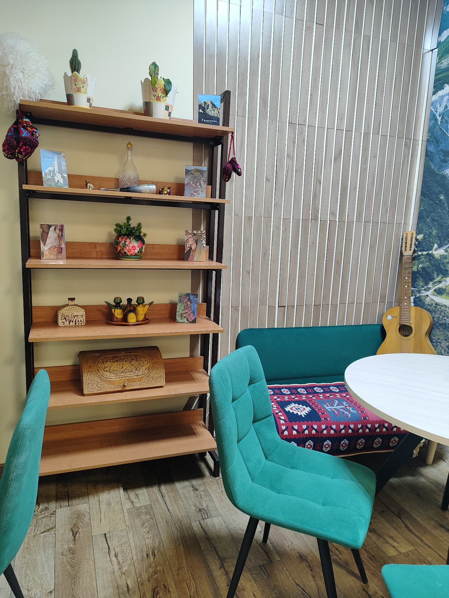 This image depicts a cozy indoor seating area, likely a lounge, cafe, or waiting room, located in Shchyolkovo, Russia. The scene is devoid of people or active events, creating a tranquil atmosphere.
The prominent feature on the left is a tall, five-shelf wooden bookcase with a black metal frame, filled with various decorative objects.
* The top shelf holds a fluffy white decorative item, two potted plants (one cactus), and two small books, one featuring the Cyrillic text "Гамустель" (Gamustel).
* The second shelf displays knitted colorful ornaments hanging from the sides, a glass decanter, small figurines, and two more small books with "Кахиб" (Kakhib) and "Ухул" (Ukhul) visible on their covers.
* The third shelf has framed artworks or cards and a potted plant with vibrant pink and red flowers.
* The fourth shelf features a decorative house-shaped bottle and a set of three cruets for oil, salt, and vinegar (words "Salt" and "Vinegar" are partly visible), alongside another artwork.
* The bottom shelf is adorned with a large, intricately carved wooden box.
To the right, a seating arrangement includes a dark teal upholstered sofa bench, accented with a patterned cushion featuring red, blue, and white geometric designs. A round white table stands in front of the sofa, flanked by two matching teal upholstered chairs with black metal legs. An acoustic guitar leans casually against the wall next to the sofa.
The background wall behind the sofa is covered in vertical light wood slats, transitioning to a large wall mural depicting a green mountainous landscape on the far right. The floor throughout the area is light-toned wooden laminate. The overall ambiance suggests a comfortable and culturally enriched space, with the visible texts potentially indicating connections to regional locations in Russia or the Caucasus.