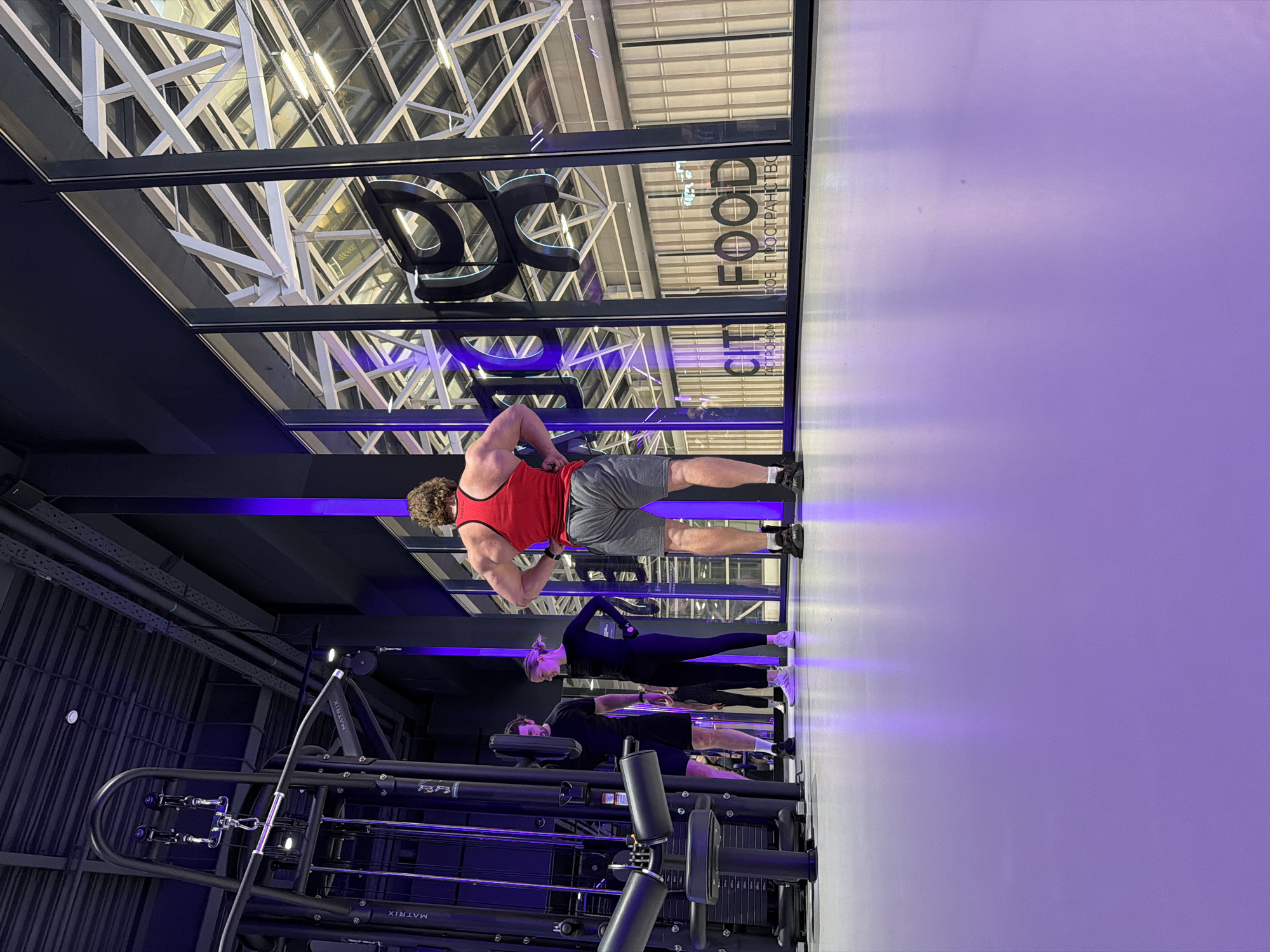 The image captures an interior scene, possibly a fitness center or gym in Saint Petersburg, Russia, based on the Cyrillic text "ЕСТРОНОМ" visible in the reflection of a window.
Three individuals are present. The closest to the camera is a man lying on an exercise bench, presumably working out. Behind him stands a woman dressed in black athletic wear. In the foreground, a muscular man in a red tank top and gray shorts is seen standing with his hands on his hips, potentially between sets of an exercise.
Gym equipment is visible, including a "Matrix" branded machine, weights, and cables, indicating the setting is a fitness center.
The interior design includes a modern architectural style with exposed structural elements and a glass ceiling. The lighting scheme is distinct, featuring blue neon lighting that adds to the contemporary ambiance of the place. The walls are painted a pale lavender color.