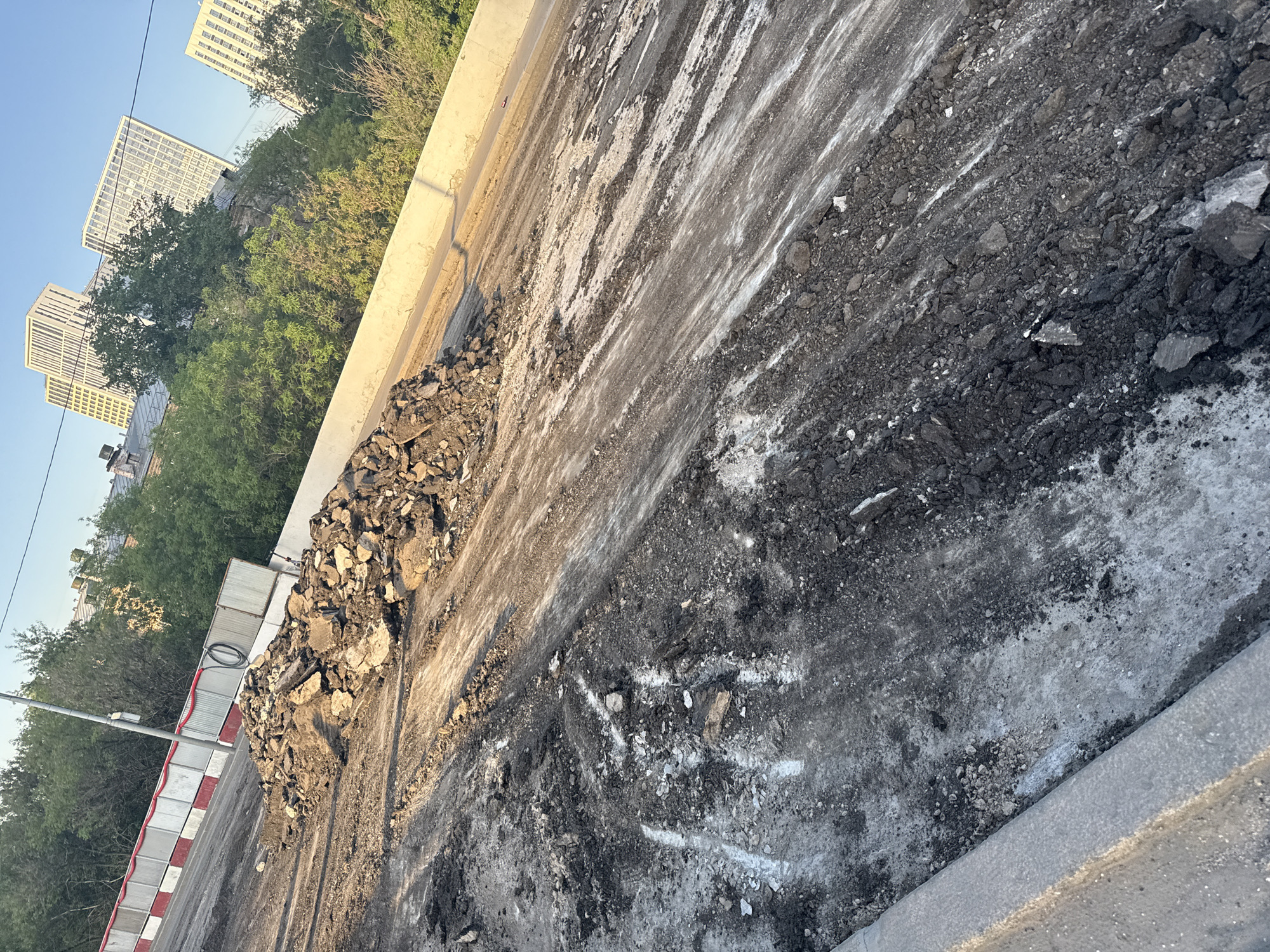 The image shows a road under construction in Khimki, Russia. Piles of broken asphalt and dirt are heaped along the edge of the road next to a gray concrete barrier. White lines are visible painted on the road surface, partially obscured by dirt and debris. Construction barriers with red and white stripes are seen in the background. Trees and tall buildings are also visible in the background, indicating an urban setting. The sky is clear and blue. No people are visible in the image.