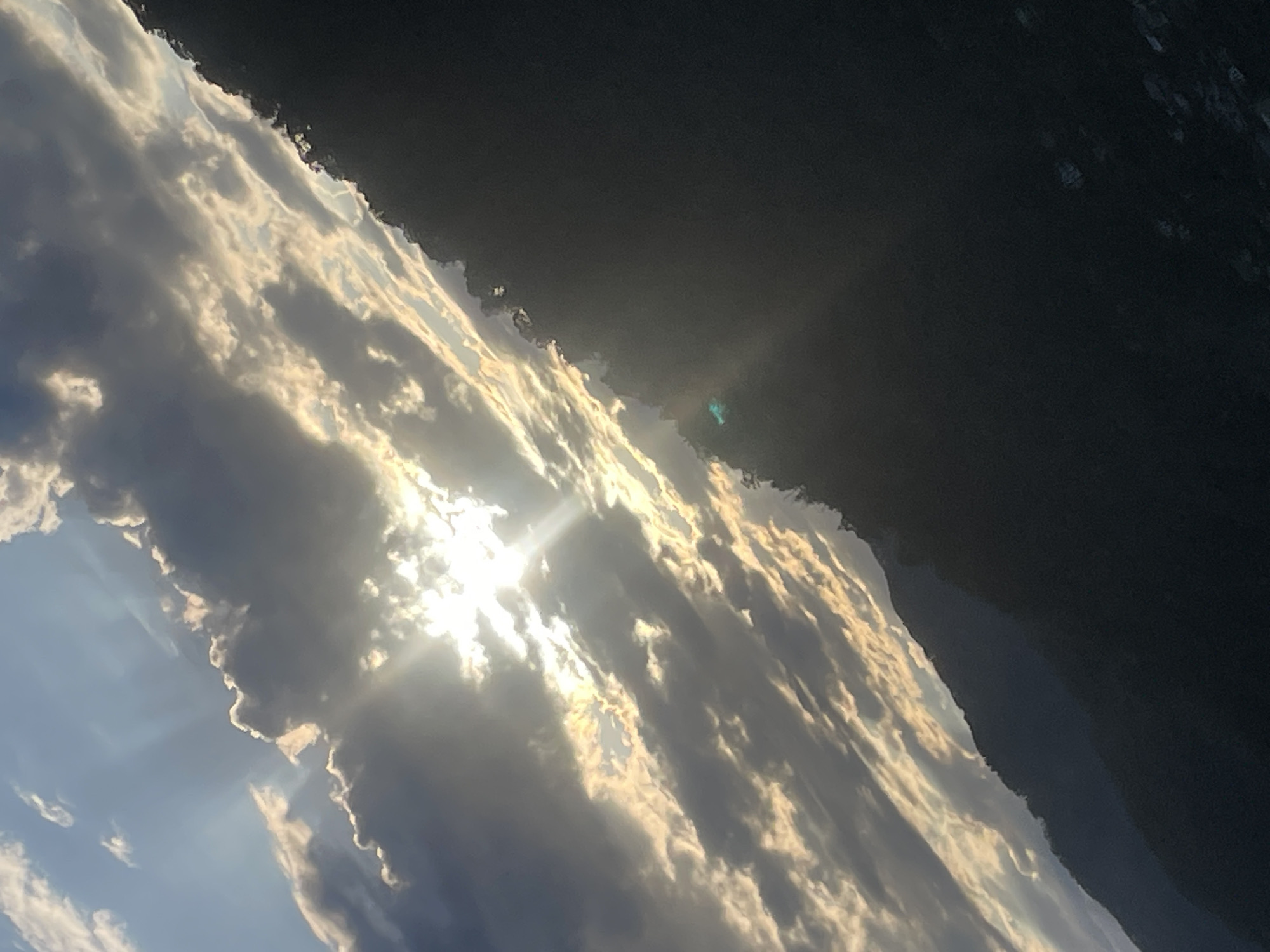 The image displays a dramatic outdoor scene, primarily featuring a dynamic sky and a dark landmass, oriented diagonally. The lower-left and central areas are dominated by an expansive cloudscape. Bright sunlight penetrates through gaps in the dense, layered clouds, casting strong, visible rays of light. The clouds themselves exhibit a range of tones, from deep grays to brilliant whites and golden highlights along their illuminated edges, suggesting either early morning or late afternoon light. The sun's intense glare is visible through an opening in the clouds.
A distinct blue-green lens flare is discernible in the darker upper-right section of the image, near the diagonal horizon line. This upper-right portion is occupied by a dark, silhouetted landform, likely mountains or hills, which forms a stark, angular boundary against the bright sky. The landmass is largely indistinct due to its shadow.
There are no people, specific objects, or visible text within the frame. The scene captures a moment of intense natural beauty, emphasizing the vastness and atmospheric conditions of the sky. The location context for this photograph is Baranovka, Russia.