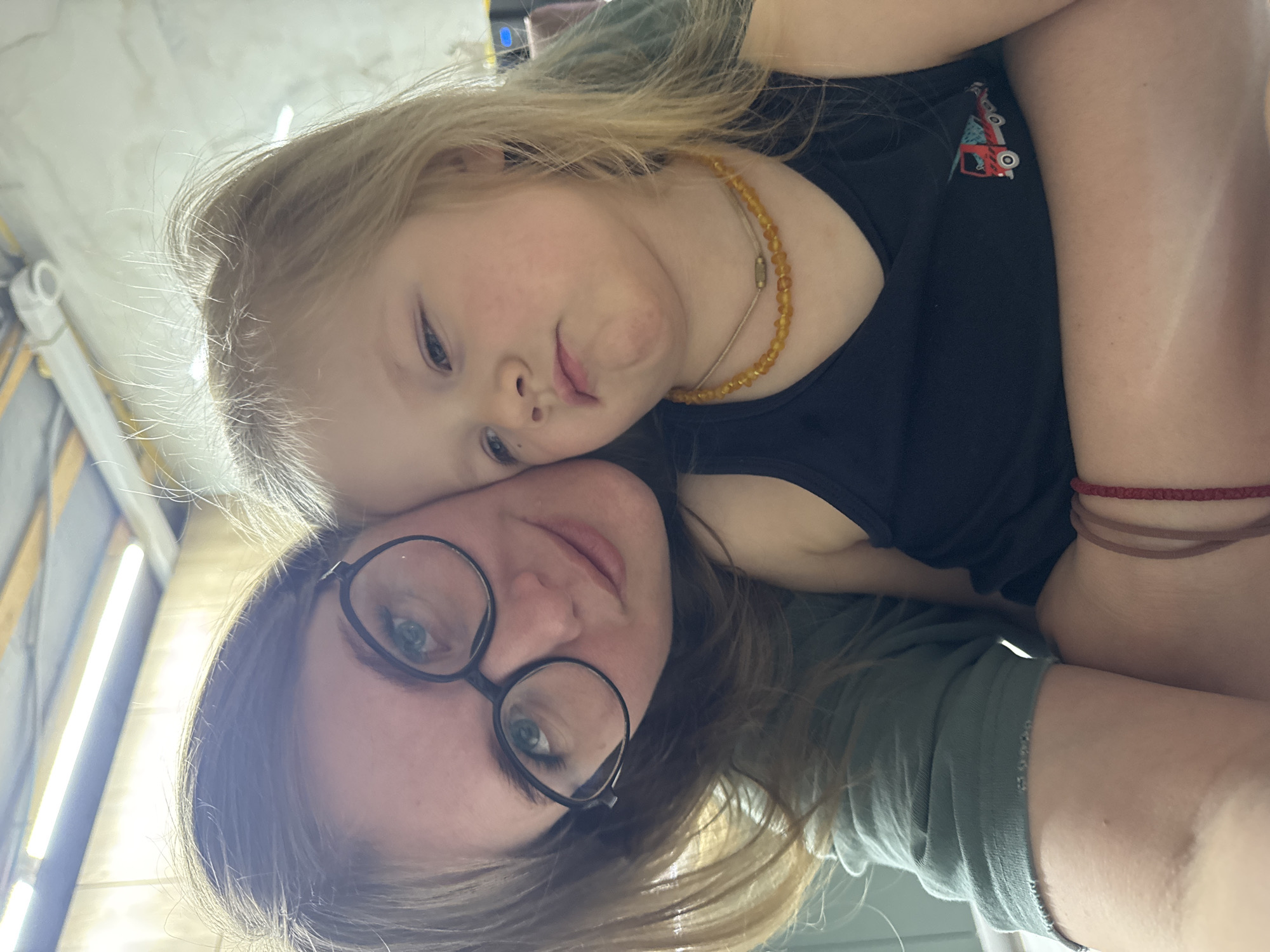 A selfie shows a woman and a child in an indoor location. The woman has brown hair, wears glasses, and a green shirt. The child has blonde hair, a black shirt, and an amber necklace. The location is possibly in Ферма, Russia.