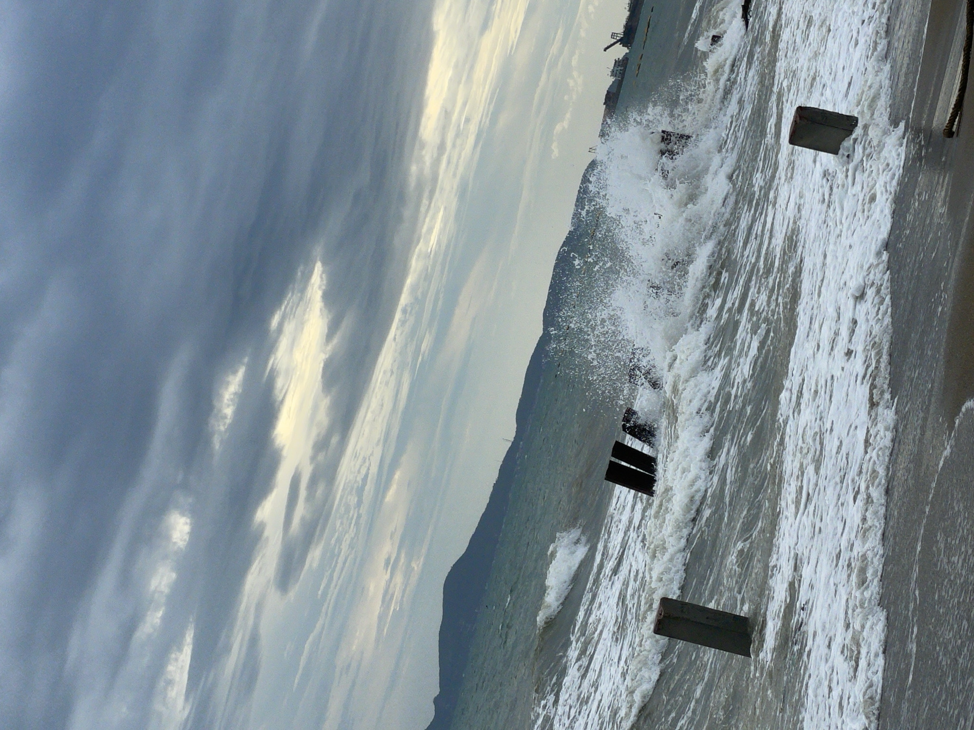 The image shows a stormy sea with large waves crashing near a coastline. The sky is overcast with dramatic clouds, and a distant mountain range is visible. In the foreground, several weathered, dark wooden posts are partially submerged in the rough water, serving as remnants of a structure or breakwater. One concrete post stands alone to the left. To the right, out at sea, a cargo ship and a crane are visible, suggesting a port or industrial area. The scene depicts a turbulent natural environment with signs of human activity. This scene is not identifiable as being in Pattaya City, Thailand.