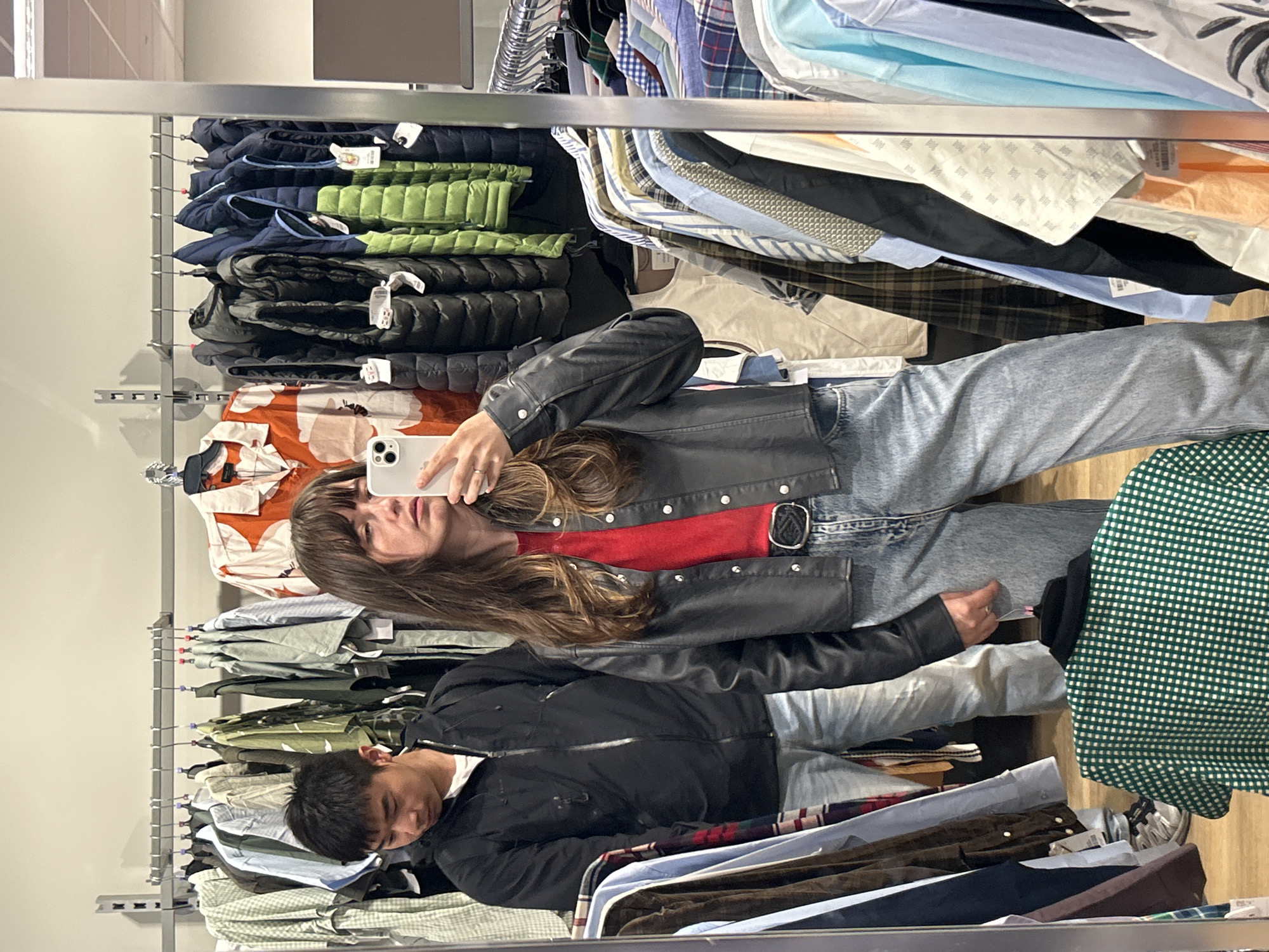 In the city of City of Edinburgh, United Kingdom, a full-length mirror reflects a young woman and a young man standing in a clothing store. The woman is in the foreground, taking a selfie with her smartphone. She has long brown hair and is wearing a red t-shirt, a black leather jacket, and light-wash denim jeans with a black belt. The man, partially visible behind her, is wearing a black zip-up jacket and jeans. They are surrounded by racks of clothing, including shirts, jackets, and vests, suggesting a second-hand or vintage clothing store environment. The background reveals more clothing on display and store fixtures, with the overall lighting indicating an indoor retail setting.