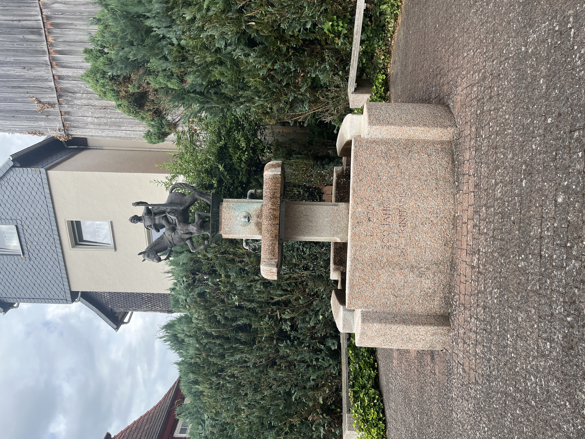 The image shows a bronze sculpture of two figures on horseback, mounted on a granite fountain. The sculpture depicts one figure appearing to lead, with the other figure following closely behind. The fountain itself is constructed of speckled pink and grey granite, with carved basins. Inscribed on the granite base of the fountain is text, which appears to read "A.D. 2004 500 JAHRE ST. TRINITAIS". The fountain is situated outdoors, with a backdrop of lush green trees and hedges. To the left of the fountain, a building with a grey, tiled roof is visible, suggesting an urban or suburban setting. The sky is overcast, indicating a cloudy day. The scene is calm and appears to be a public space, possibly a park or a square in Neustadt, Germany.
