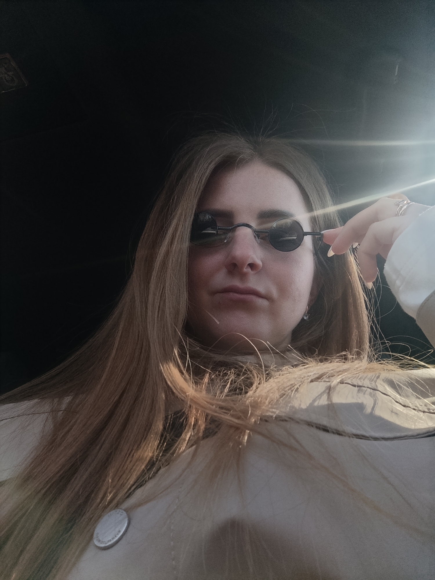 A low-angle, medium shot depicts a light-skinned woman with long, light-brown hair, wearing small, round, black sunglasses. She is holding the right side of her sunglasses with her left hand, revealing manicured fingernails. She is wearing a light-colored collared shirt with a button on the lower left. The background is dark, but there is a bright light source behind her to the right. The location is in the city of Unknown, Unknown.