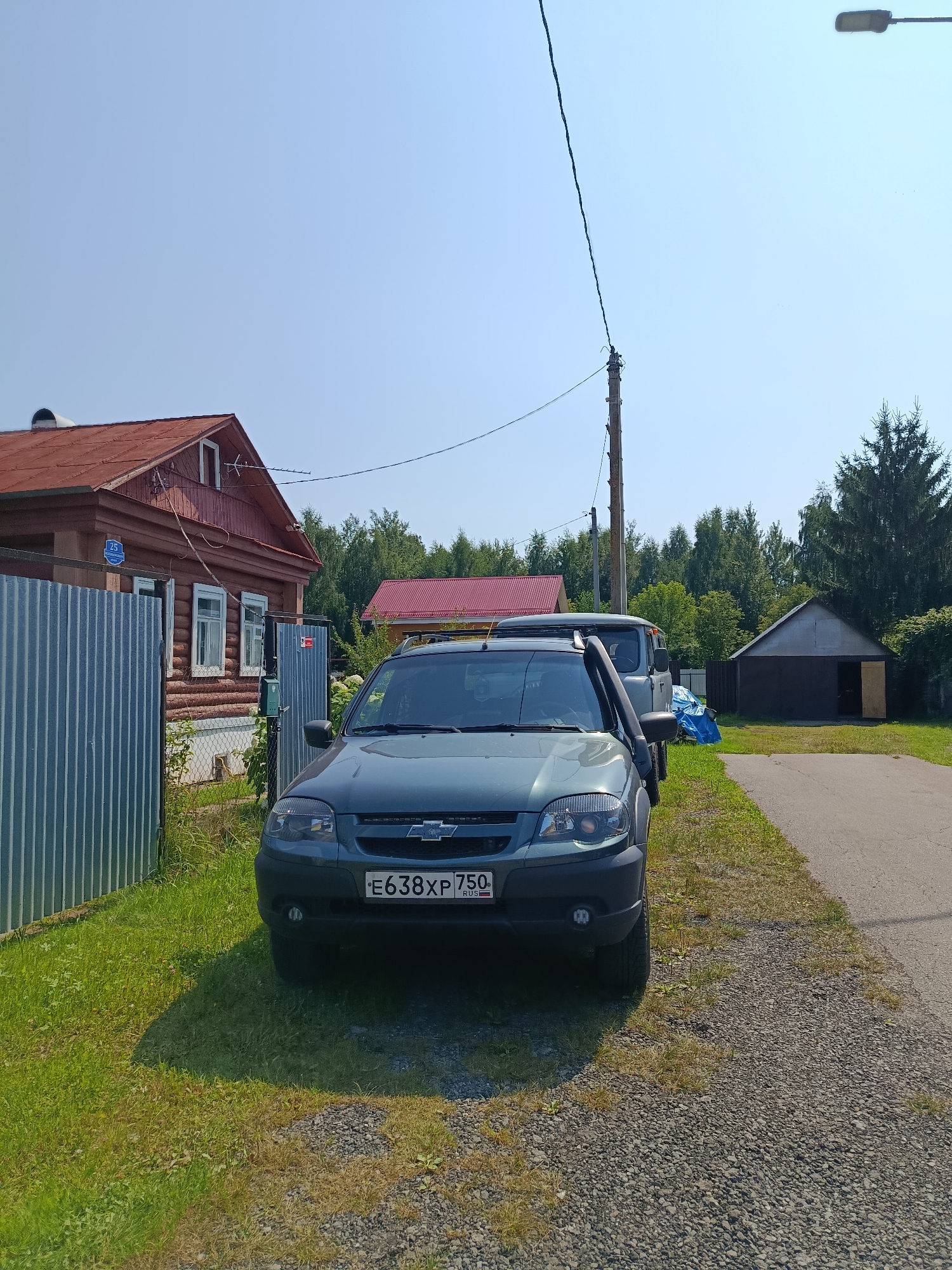 The image depicts a street scene in the city of Vereya, Russia, under a clear blue sky. In the foreground, a dark green Chevrolet Niva SUV is parked on the right side of a gravel and asphalt road. The car's license plate reads "E638XP 750". To the left of the car, a corrugated metal fence encloses a grassy yard with a traditional wooden house. The house has a red roof, white-trimmed windows, and a sign with the number "25" and Cyrillic lettering. Behind the house, more trees and another building with a red roof are visible. Further down the road, a detached garage with brown doors and an open entrance stands among trees. A wooden utility pole with electrical wires runs vertically through the scene. A blue tarp is visible in the background near the garage. The environment appears to be rural or suburban, with a focus on residential structures and vehicles.