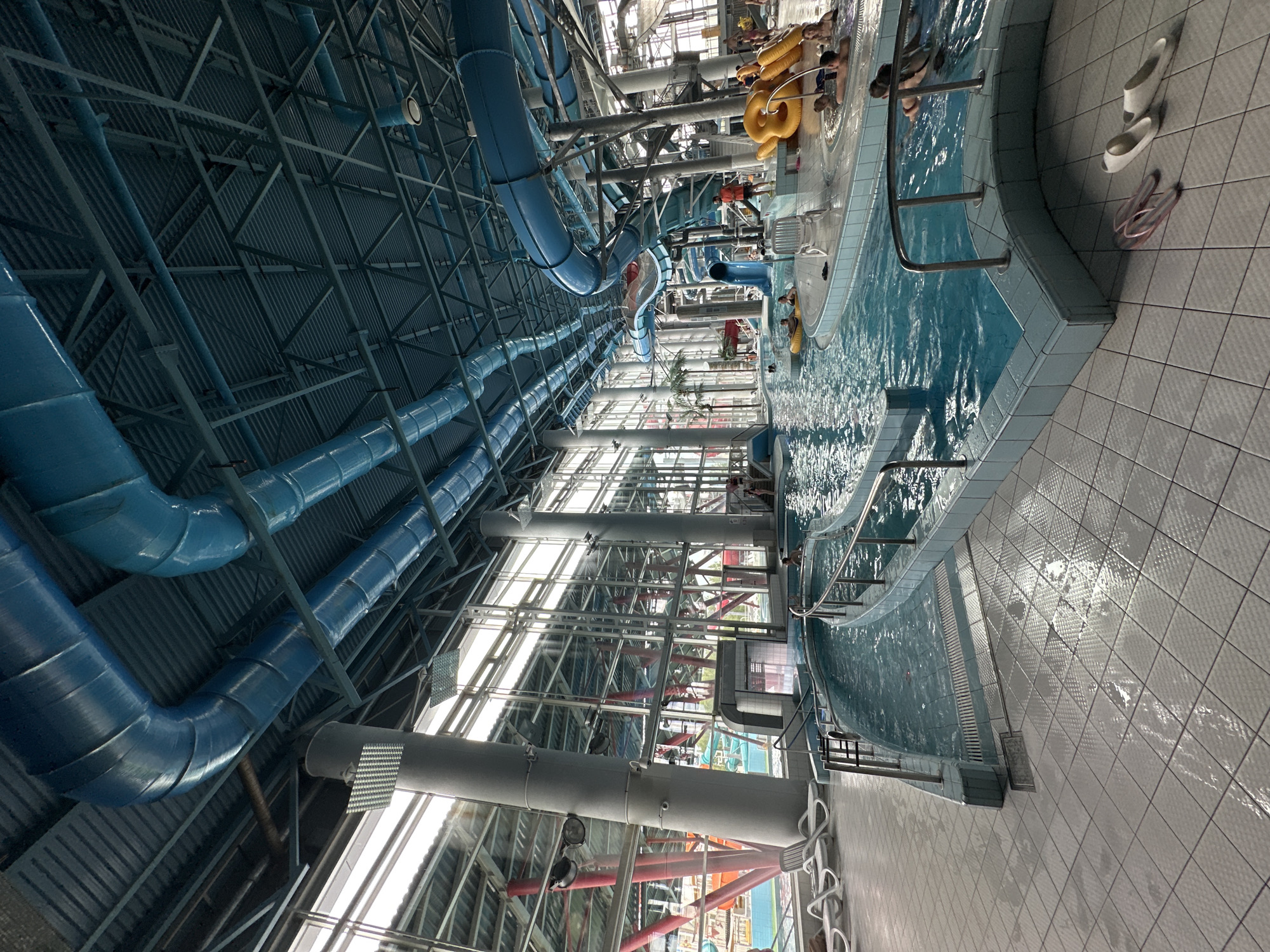 The image depicts the indoor water park "11 Vody" (11 Waters) located in Minsk, Belarus. The scene features a large swimming pool area with several blue water slides visible, winding from the ceiling down to the pool. Natural light streams in from large windows along the side of the building, illuminating the blue-tiled interior and the water. A lazy river or a shallow wading pool is in the foreground, with people relaxing in the water. White lounge chairs are positioned near the pool on the tiled floor. Several people are seen in and around the water, some on inflatable tubes. The ceiling is a complex structure of metal beams painted blue, with ventilation pipes also prominently displayed. Two pairs of flip-flops, one pink and one white, are placed on the tiled floor in the foreground. The overall atmosphere is that of a recreational water park on a bright day.