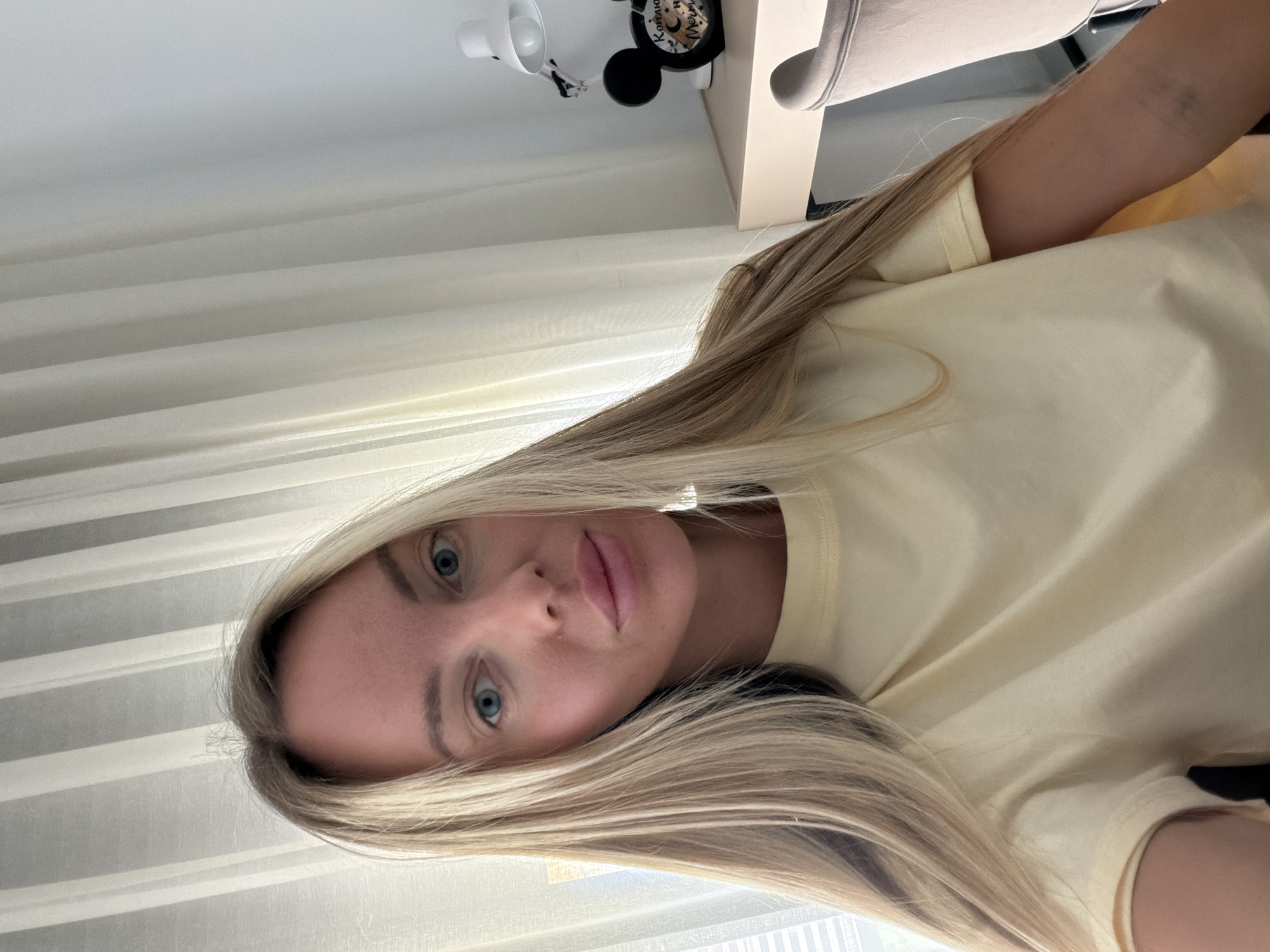 A fair-skinned woman with long blonde hair and blue eyes is pictured in a selfie taken indoors. She is wearing a light yellow t-shirt. The background consists of sheer white curtains and a desk with a lamp and a Mickey Mouse-shaped clock. The woman appears to be at home or in a hotel room in Finestrat, Spain, based on the context provided. The lighting is natural, coming from a window behind the curtains. The overall scene is casual and domestic.