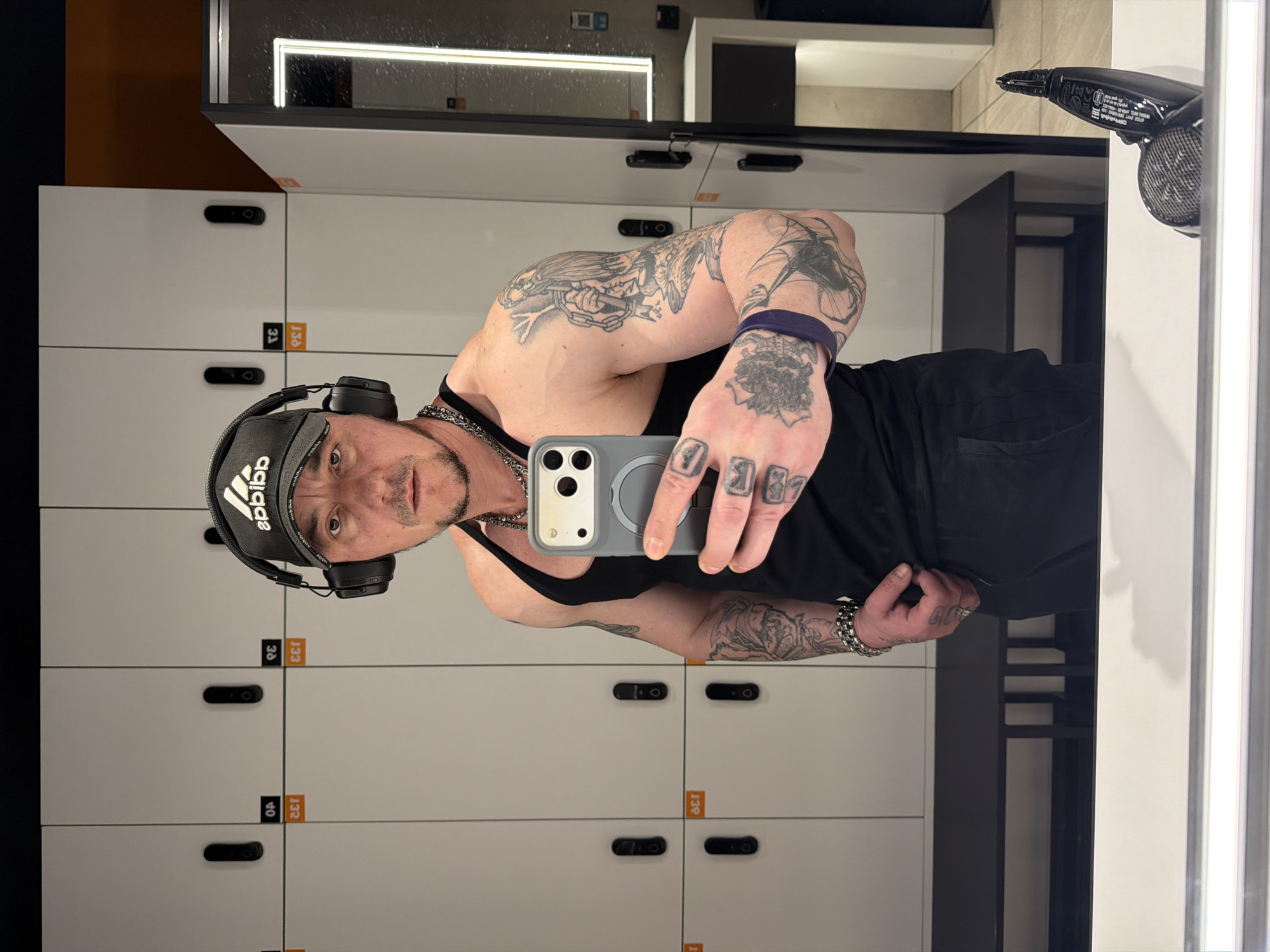A man is taking a selfie in a locker room. He is wearing a black Adidas cap, headphones, and a black tank top. He has numerous tattoos on his arms and hands. The background shows rows of white lockers with black handles. A hair dryer is visible on the right side of the image. There is no evidence to suggest the image was taken in Moscow, Russia.