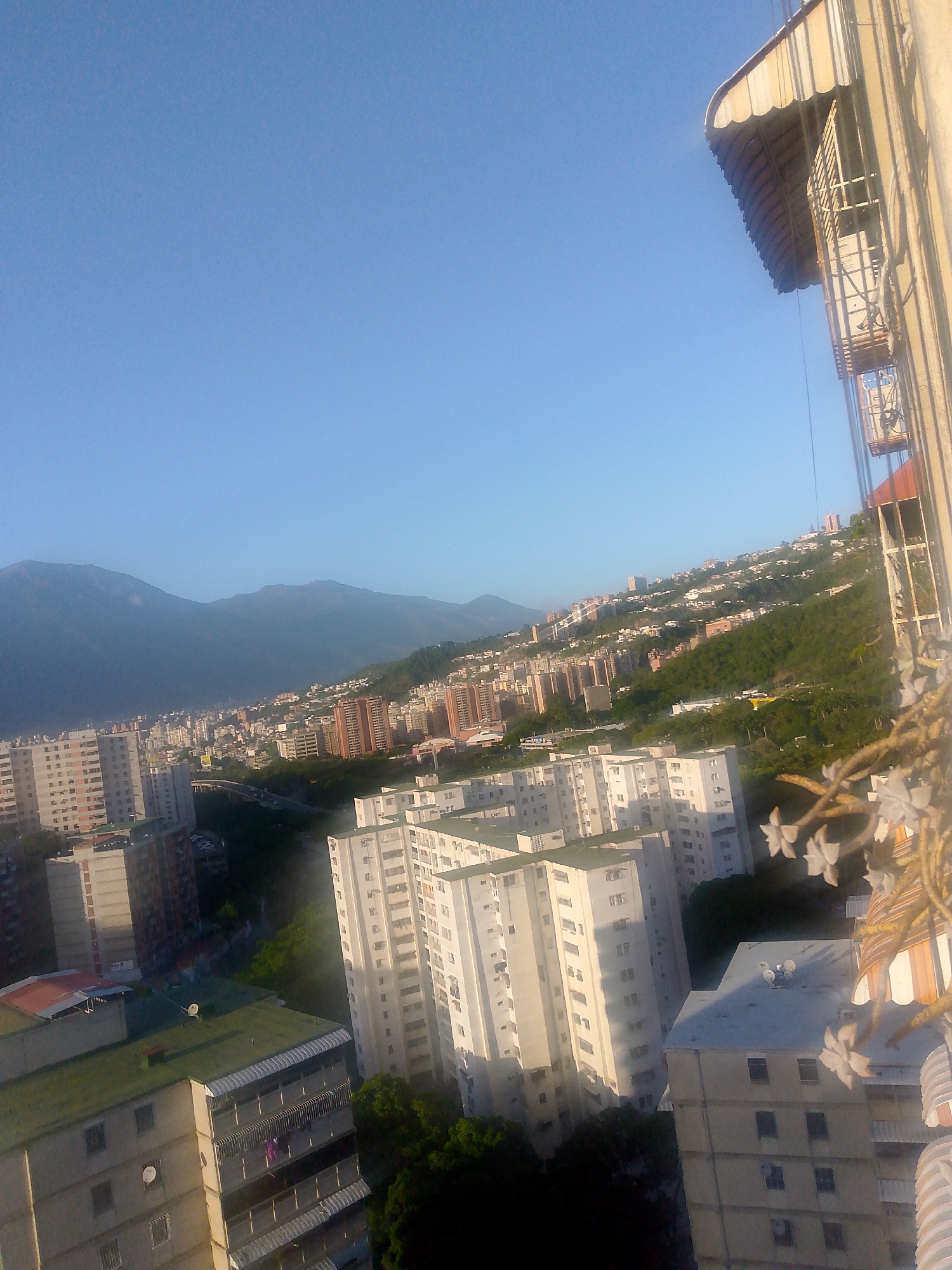 This is a high-angle, wide-shot photograph overlooking a densely populated urban area in Caracas, Venezuela, likely taken from a building in the late afternoon or early morning given the clear sky and long shadows. The foreground features a portion of a building's exterior with a cream-colored awning and a partially visible metal structure, possibly a lift or scaffolding, with some floral elements in the lower right corner.
The midground and background are dominated by a vast cityscape with numerous multi-story apartment buildings of varying heights and architectural styles. Many buildings are white or light-colored, featuring numerous windows. Some structures are clustered closely together, while others are spread out across the landscape. The city extends up a steep, green hillside, indicating a mountainous topography. The distant background shows imposing, hazy mountains that form a natural boundary to the urban sprawl.
In the lower sections of the image, darker green foliage from trees is visible amongst the buildings, suggesting pockets of vegetation within the city. The overall impression is one of a sprawling, vertical city nestled against a mountainous backdrop. There are no discernible people, vehicles, or specific activities captured in the frame.
