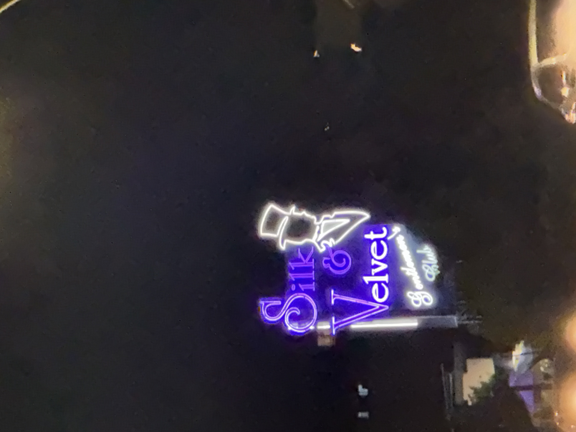 The image captures a neon sign that reads "Silk & Velvet Gentlemen's Club" in the city of Mouttagiaka Community, Cyprus. The sign is illuminated in purple and white, with the silhouette of a man in a top hat integrated into the design. The background is dark, suggesting nighttime, and there are some faint lights and structures visible in the lower part of the image, possibly indicating the surrounding urban environment. A vehicle is partially visible in the bottom right corner.