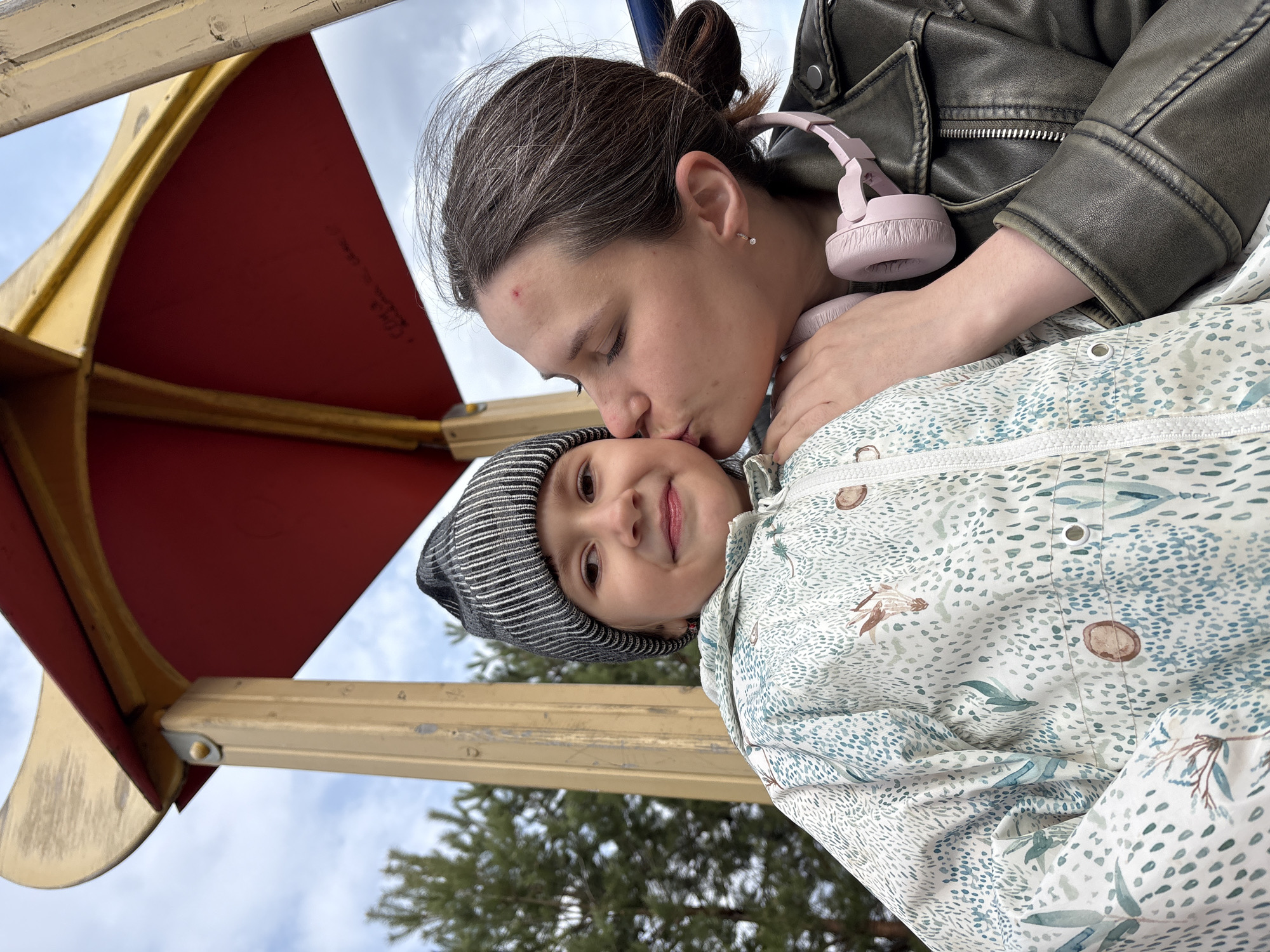 The image captures an affectionate moment between a woman and a young child, tilted on its side, likely taken from a low angle outdoors in Chaikovsky, Russia. The background features a cloudy sky and the tops of pine trees, along with parts of yellow and red playground equipment.
The woman, with dark hair pulled back, wears a dark, possibly olive green, leather-style jacket. Pink wireless over-ear headphones are around her neck. She is leaning down to gently kiss the young child on the forehead or temple. A small red mark is visible on her forehead, and she has a small, sparkling earring in her right ear.
The child, appearing to be a toddler or preschooler, wears a gray and black striped beanie and a light-colored puffy jacket adorned with a pattern of green and blue dots, leaves, and small brown shapes. The child looks towards the camera with a slight, contented smile.
The scene suggests an intimate and tender interaction between the two, likely a mother and child, enjoying an outing on an overcast day. Faint, illegible white markings are present on the red surface of the playground structure.