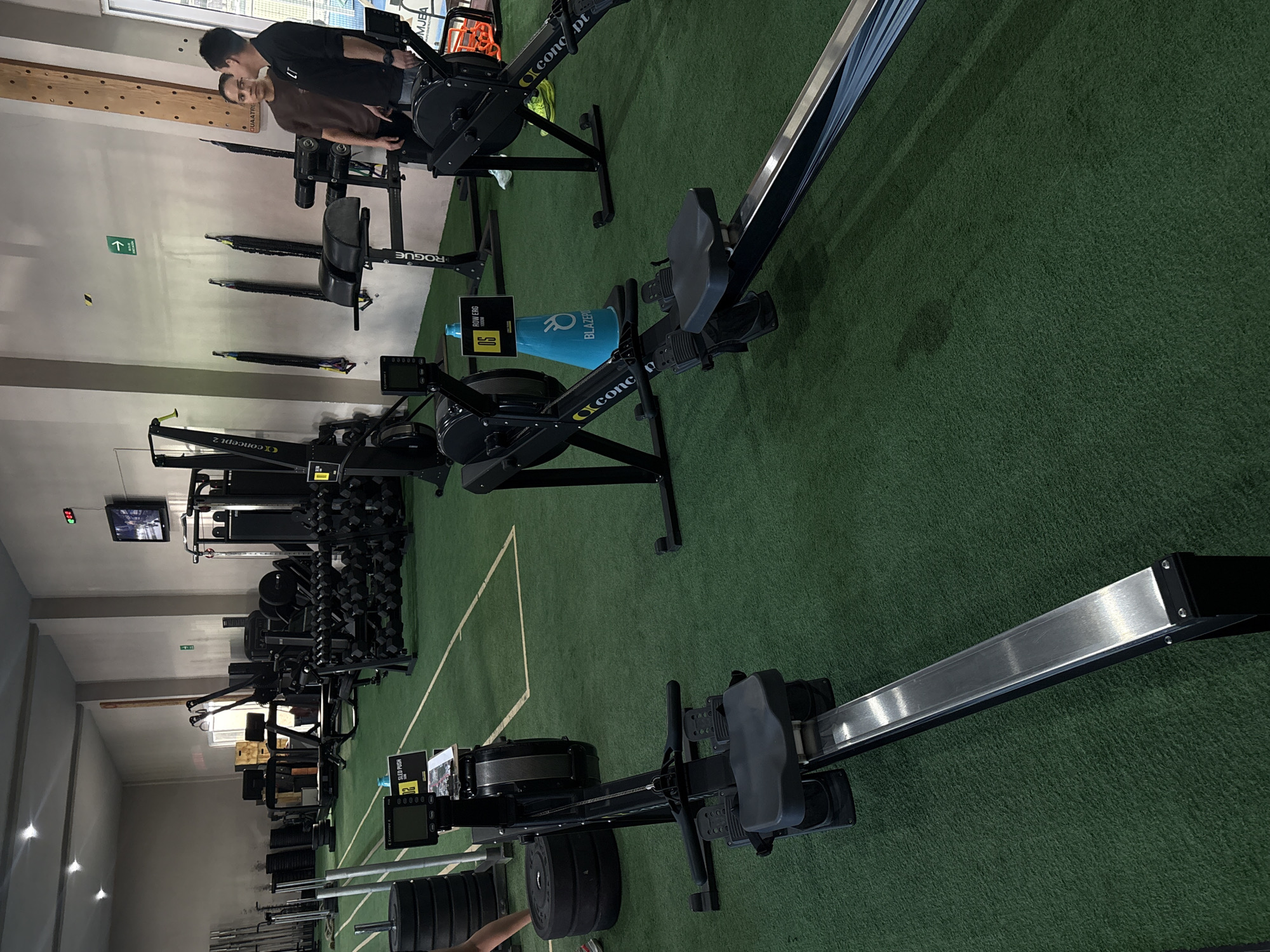 The image depicts the interior of a gym, possibly located in San Bernardino Tlaxcalancingo, Mexico, although there is no definitive evidence of the city. The gym features multiple Concept2 rowing machines arranged on a green turf floor. Other gym equipment, including weight racks with dumbbells and a cable machine, are visible in the background. Two individuals are seen in the upper left corner of the frame near the entrance, possibly engaged in a workout. An emergency exit sign is mounted on the wall. A water bottle with the label "Blazer" is seen next to one of the rowing machines.