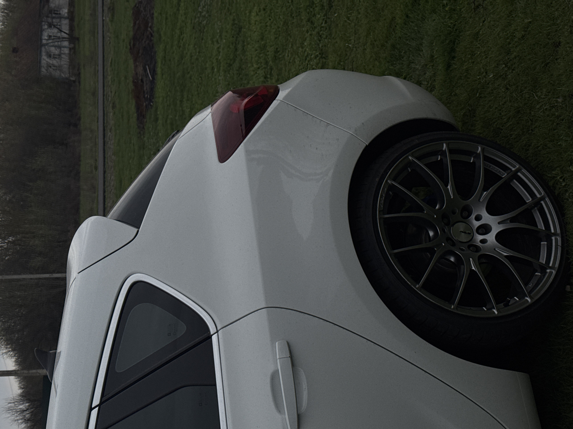 The image shows the rear quarter of a white car parked on a grassy area. The car features a dark-colored wheel with a multi-spoke design. A red tail light is visible. The background consists of grass and what appears to be trees and a building in the distance. Without further data, it is not possible to confirm that the picture was taken in Semeno-Aleksandrovka, Russia.