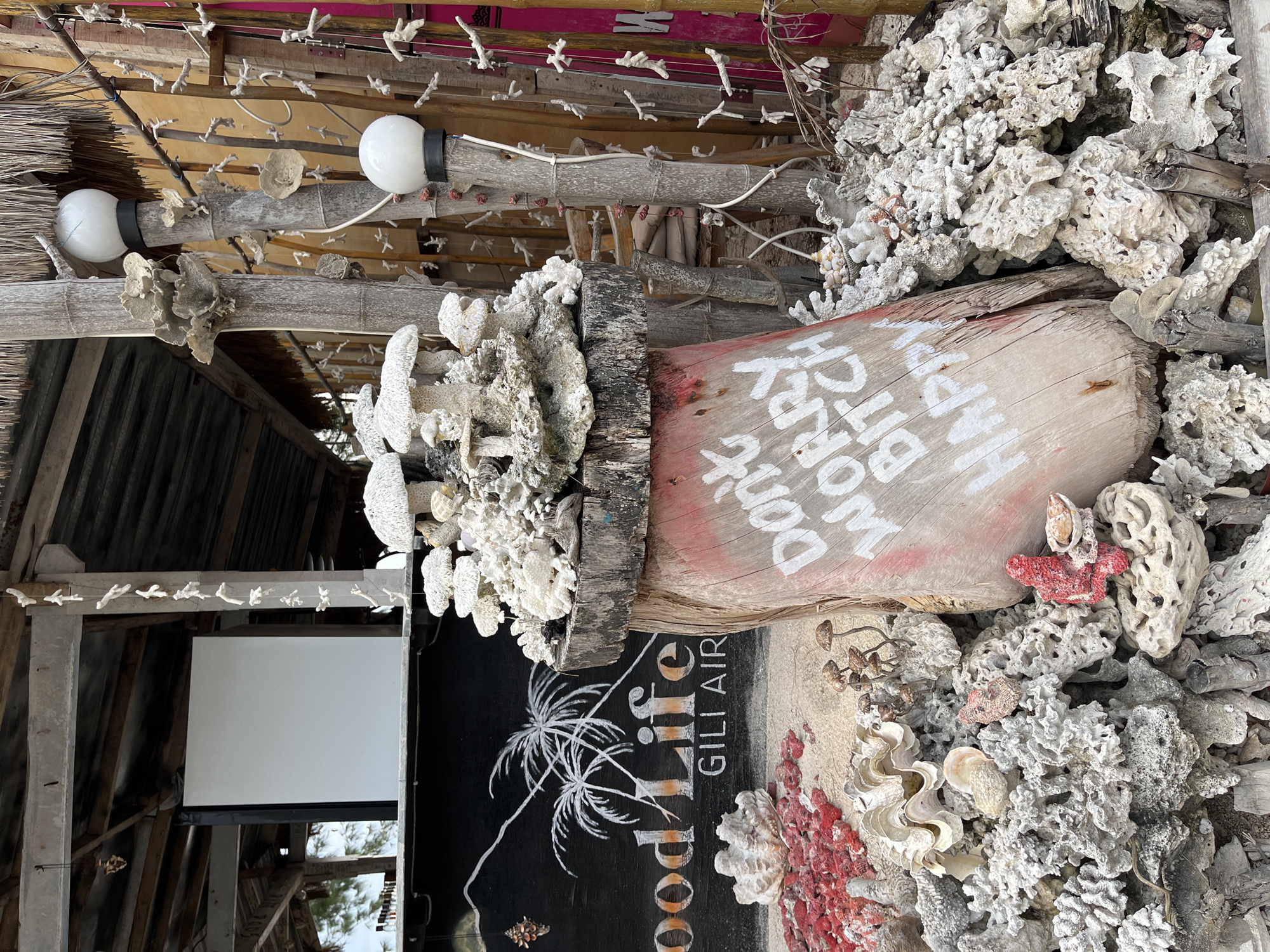 The image shows a decorative structure in Unknown, Indonesia. A piece of weathered wood has the phrase "Don't Worry Be Happy Bitch" painted on it in white lettering. The wood is surrounded by pieces of coral and shells. A black sign with a palm tree design and the text "Goodlife GILI AIR" is also visible. The structure includes light bulbs and various decorative elements made of wood and coral. The overall environment appears to be a beachside or tropical setting.
