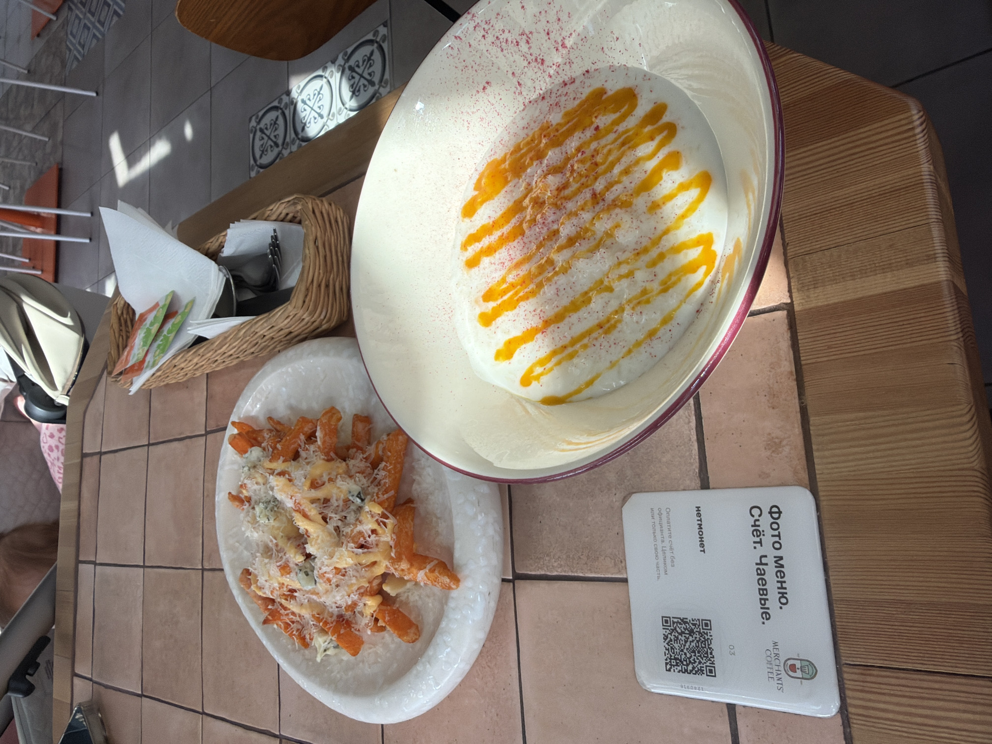 This image shows a table with food and a card. The table appears to be inside a restaurant or cafe. On the table are two plates with food. One plate contains what appears to be a sweet dish with a white base and orange drizzle, topped with small red sprinkles. The other plate holds orange-colored fries or sweet potato fries topped with cheese and sauce. Beside the plates is a white card with text in Russian, including the words "Фото меню. Счёт. Чаевые," which translates to "Photo menu. Bill. Tips". The card also has a QR code and the logo for "MERCHANTS' COFFEE". Next to the plates is a wicker basket with napkins and cutlery. Based on the presence of Russian text and the card, the location is likely in Moscow, Russia.