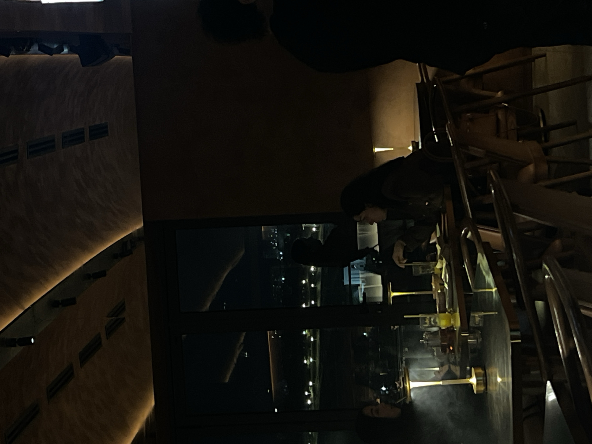 The image depicts the interior of a building, possibly a bar or restaurant, overlooking Yerevan, Armenia at night. The scene is dimly lit, with a large window providing a view of the illuminated city skyline. Silhouettes of people are visible both inside the building and reflected in the window. Inside, several people are standing around a bar or counter, while chairs and a railing are also visible. The exterior view shows streetlights and building lights of Yerevan.
