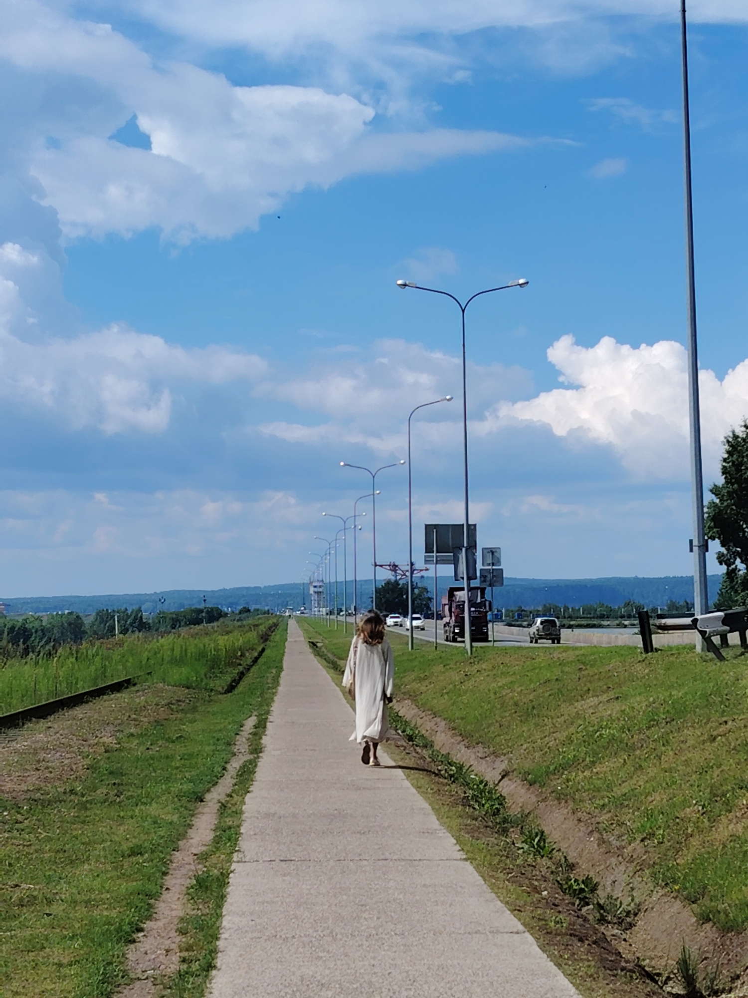 A full-shot image captured from behind shows a person in a light-colored robe walking away from the camera on a concrete sidewalk. The sidewalk runs parallel to a grassy area with a railroad track to the left. To the right of the sidewalk is a road with several vehicles, including trucks and cars, traveling in the same direction as the person. A line of streetlights stands between the sidewalk and the road. In the distance, a city skyline is visible with a backdrop of trees and hills under a bright blue sky with scattered clouds. The scene appears to be the outskirts of the city of Chaikovsky, Russia, specifically along a road that leads into the city. The lighting suggests it is daytime.