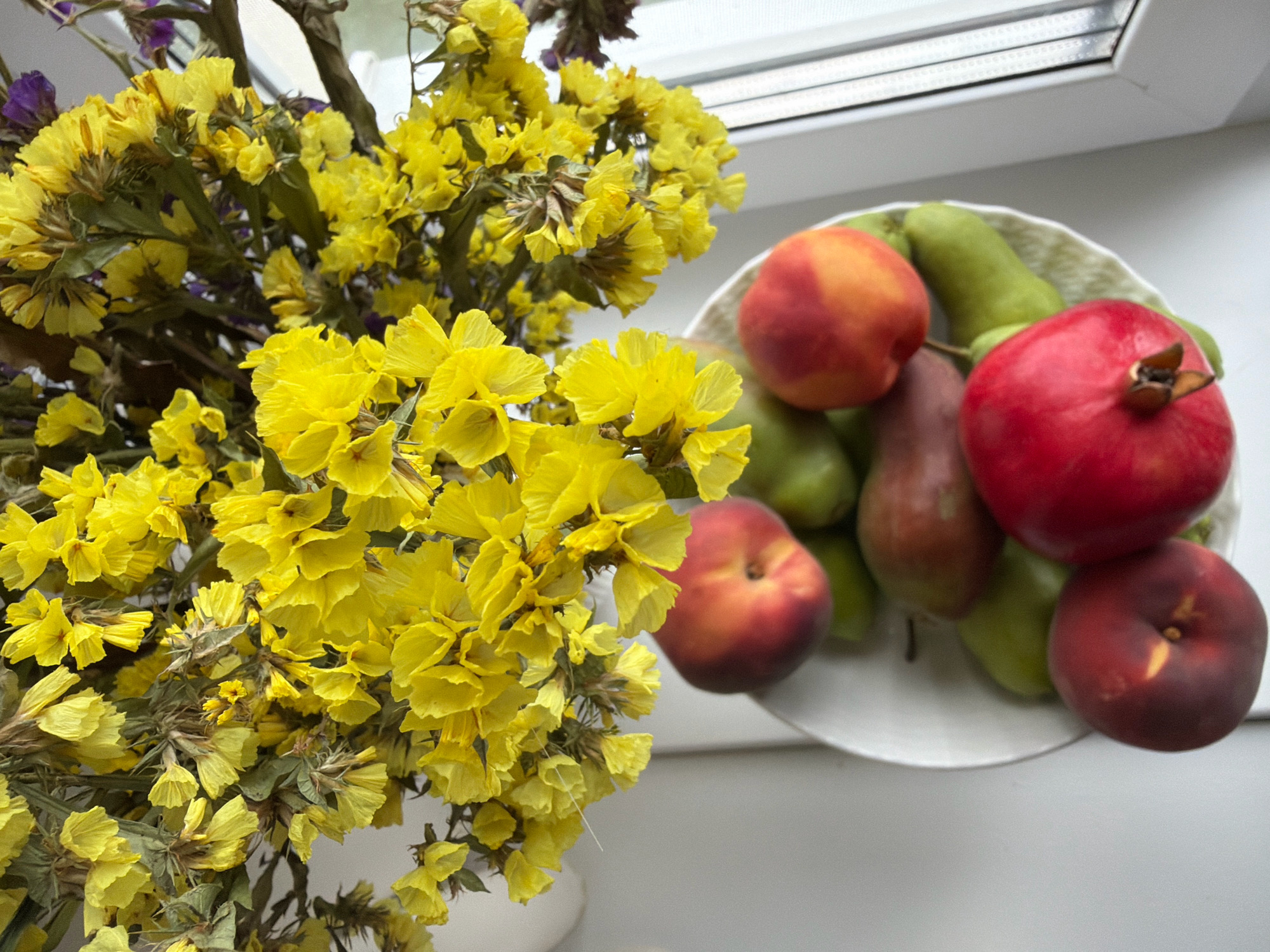 A close-up, high-angle shot shows a bowl of fruit and a bouquet of dried yellow flowers. The fruit bowl, which is white with a textured rim, contains several items including a red pomegranate, two green pears, and two peaches, one with predominantly red and yellow coloring and the other a deep red. The dried flowers are a vibrant yellow, with delicate, intricate petals, and are arranged in a bouquet to the left of the fruit bowl. Purple flowers are also interspersed within the bouquet. The scene appears to be set on a windowsill, with the window frame and a sliver of a light-colored wall visible in the background. The image is sharp and well-lit, with the focus primarily on the fruit and flowers. The overall impression is one of a still life arrangement, possibly in a domestic setting. No people are visible in the image, and no specific location within Бабініцкі сельскі Савет, Belarus can be determined from the visual evidence.
