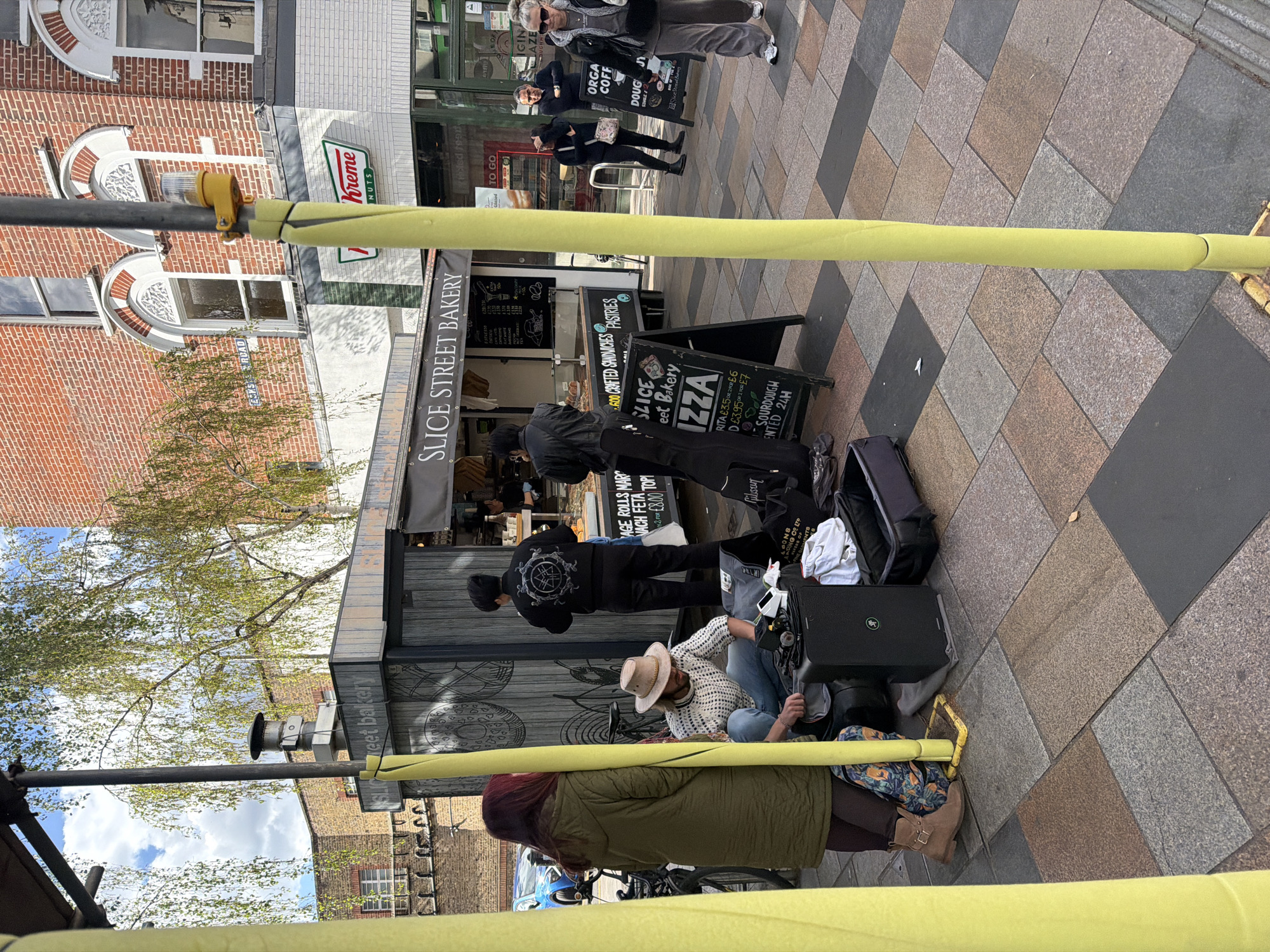 The image shows a scene on a street in what appears to be London, United Kingdom. A Slice Street Bakery is visible, with a sign advertising "Pizza" and other baked goods. There is a Krispy Kreme sign in the background as well. Several people are visible: customers at the bakery, people standing near a sandwich board, and a street performer sitting on the ground with a speaker and a guitar. Scaffolding, padded with yellow material, is in the foreground. The paving is a mix of grey, brown and black rectangular bricks. The sky is visible through some trees on the left.