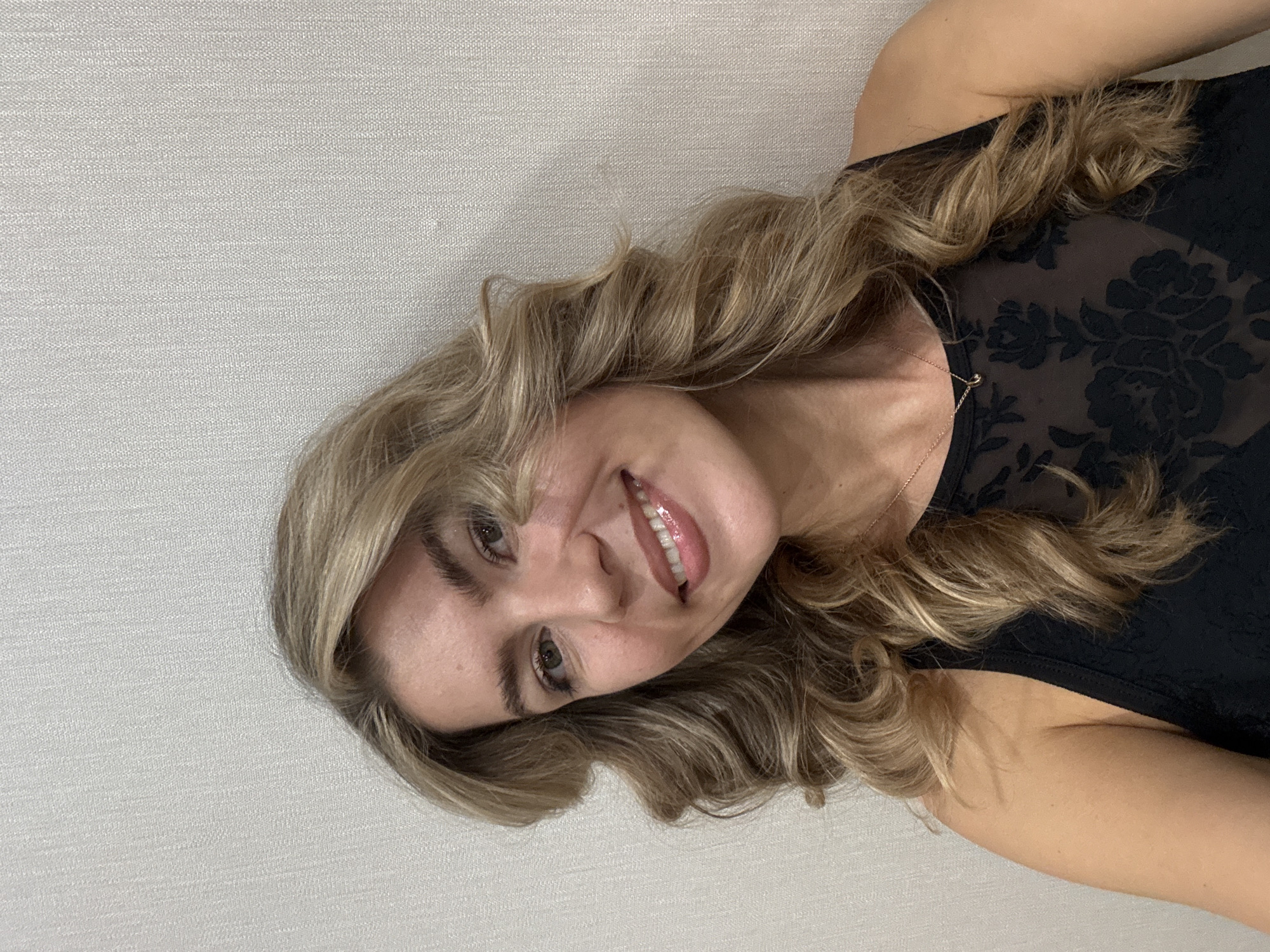 A vibrant portrait captures a smiling young woman with long, wavy, blonde and light brown hair. She is looking directly at the camera with a cheerful and friendly expression, her head tilted slightly to the side. A wide smile reveals her teeth, and her lips have a glossy finish. She has prominent eyebrows and appears to be wearing some light eye makeup.
The woman is dressed in a dark-colored top, likely black, which features a sheer or lace overlay on the upper chest and shoulder area, detailed with a subtle floral pattern. A thin, gold-toned necklace with a small, circular pendant is visible around her neck. Her hair flows around her face and shoulders, adding to the dynamic feel of the image.
The background is a plain, light-colored wall with a subtle fabric-like texture, suggesting an indoor setting. The lighting is soft and even, illuminating her face and highlighting the waves in her hair. There is no visible text in the image.