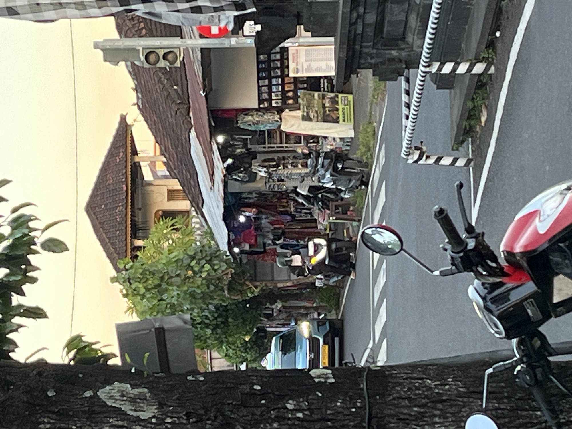 The image depicts a street scene in an urban area of Unknown, Indonesia. A motorcycle is prominently positioned in the foreground, with its handlebars and mirrors clearly visible. The street leads to a busy commercial area with shops and vendors. There's a mix of pedestrian and vehicle traffic. A building with a traditional Balinese-style roof is visible. The lighting indicates daylight. A light blue vehicle is driving on the street behind several scooter drivers. A pedestrian is passing the scooter drivers.