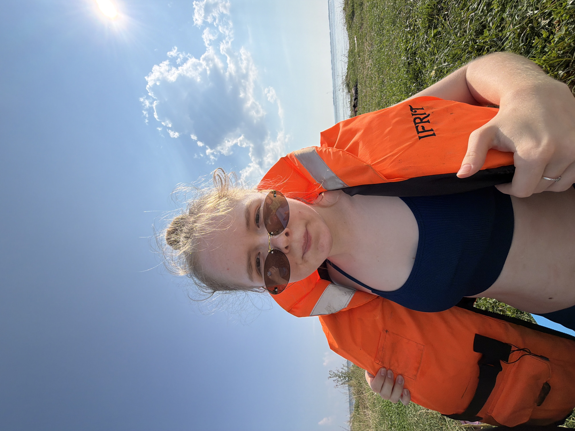 A young woman with blonde hair pulled up in a bun, wearing sunglasses and an orange life jacket over a navy blue bikini top, is pictured outdoors on a grassy area near a body of water under a clear blue sky with scattered clouds and bright sunlight. She is looking at the camera and holding the edges of the life jacket. The word "IFRIT" is visible in black lettering on the life jacket. The background shows a body of water with a faint shoreline in the distance. The overall scene suggests a sunny day by the water.