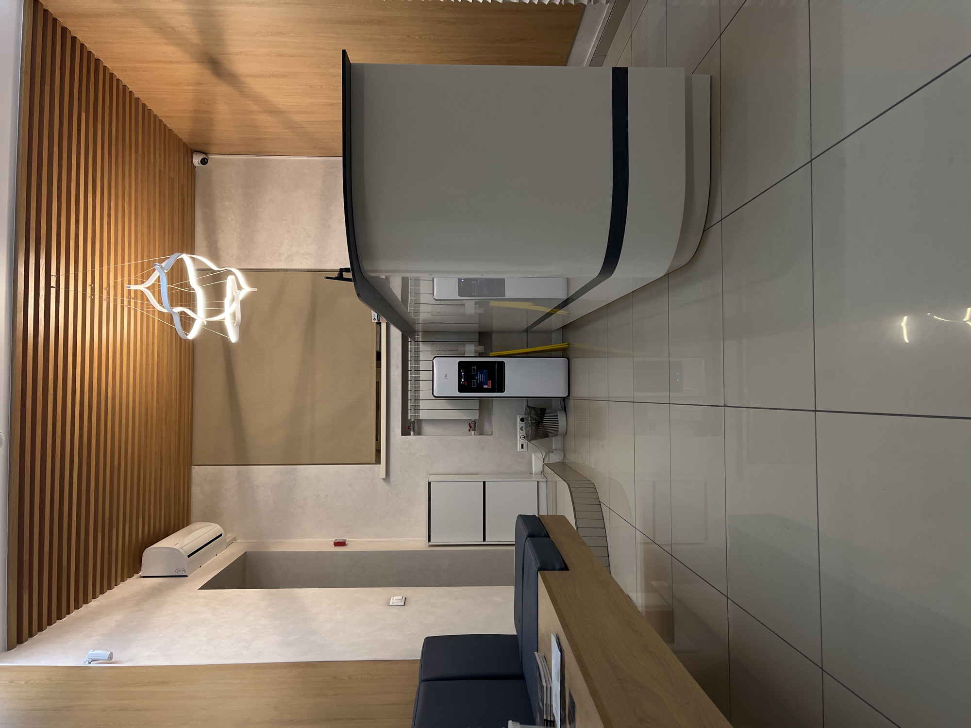 The image shows the interior of a modern building in what is likely Krasnoyarsk, Russia.
**Environment:** The space is brightly lit and appears to be a waiting area or lobby. Walls are primarily a light beige color, with one wall featuring a vertical wooden slat design. The ceiling is partially covered with wood paneling. The floor is tiled in a light color.
**Objects:**
* A large, curved, white structure (possibly a reception desk) dominates the scene. It has a black trim.
* There is a smaller machine with a screen and buttons, situated near the large curved structure.
* A white air conditioning unit is mounted on a wall.
* A modern, decorative light fixture hangs from the ceiling.
* A blue upholstered bench sits near a light wood table, suggesting a waiting area.
**Inferences:** The clean, modern design suggests a relatively new or recently renovated space, possibly a clinic, office building, or upscale service establishment. Given the limited information, it is difficult to specify the location in Krasnoyarsk.