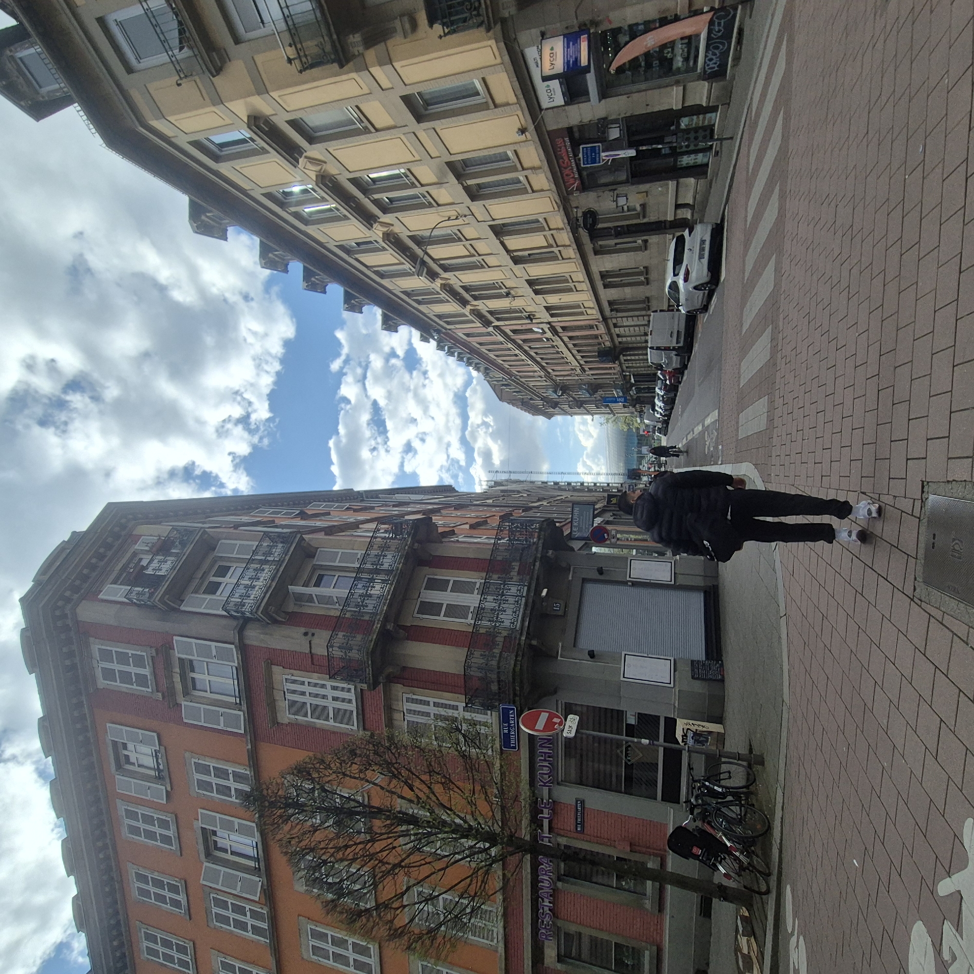 The image captures a street scene in Strasbourg, France. The perspective is slightly angled, showcasing buildings lining the street under a sky filled with clouds. A person is walking along the sidewalk paved with brick tiles. A sign on the street says "Rue Thiergarten." Another sign reads "Restaurant le Kuhn". Several cars are parked along the street. Bicycles are parked along the building near the sidewalk.
