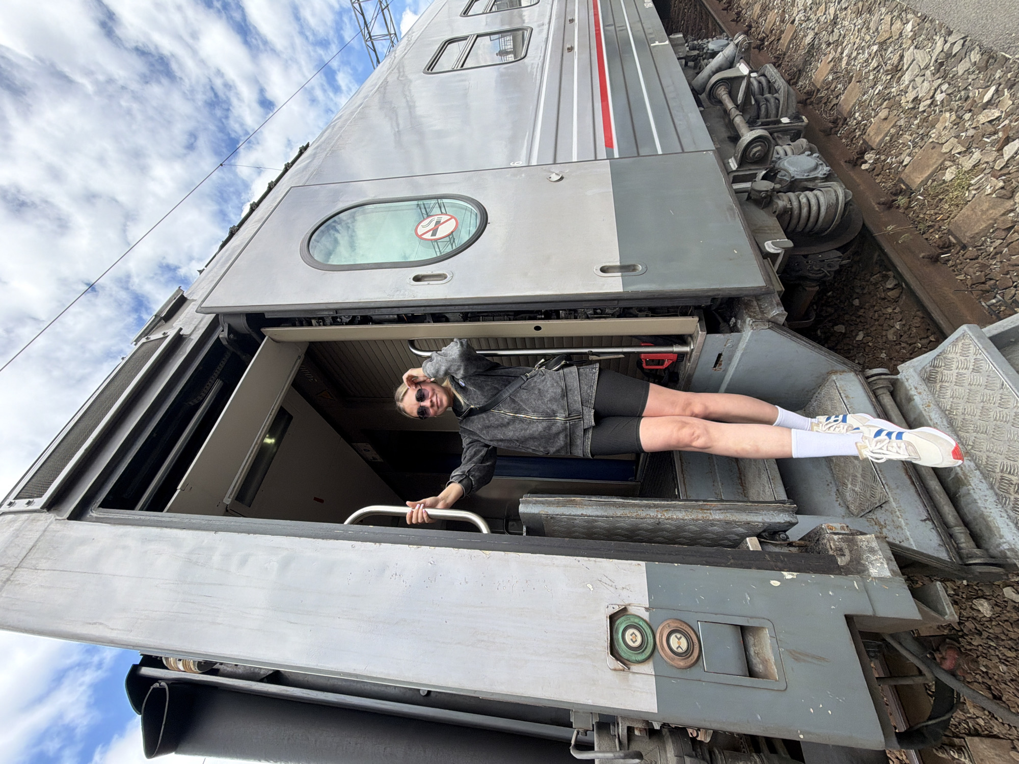 A young woman is standing in the open doorway of a train car. She is wearing black bike shorts, a gray jacket, white socks, and white sneakers with blue stripes and red accents. She is also wearing sunglasses and has her hand on a silver grab rail. The train car is gray with a red stripe and a "no smoking" sign visible on the window above the door. The train is on tracks surrounded by gravel and concrete. The sky is partly cloudy. This image was taken in Rostov-on-Don, Russia.