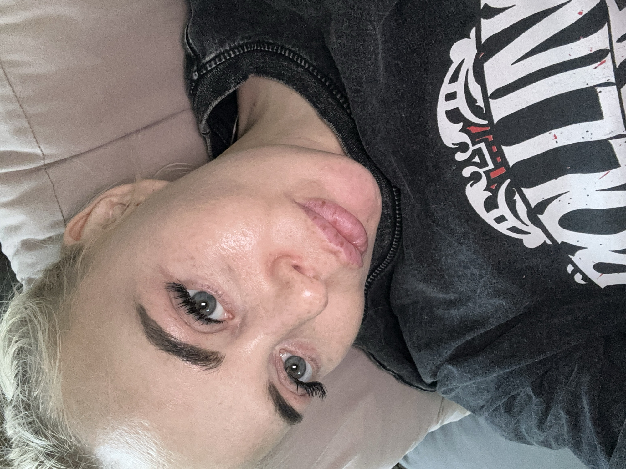 A woman with blonde hair and gray eyes is lying on a light-colored pillow or surface. She is wearing a dark gray sweatshirt with a white design on the right side of the frame. Her face is slightly angled towards the camera. The location is not discernible from the image.