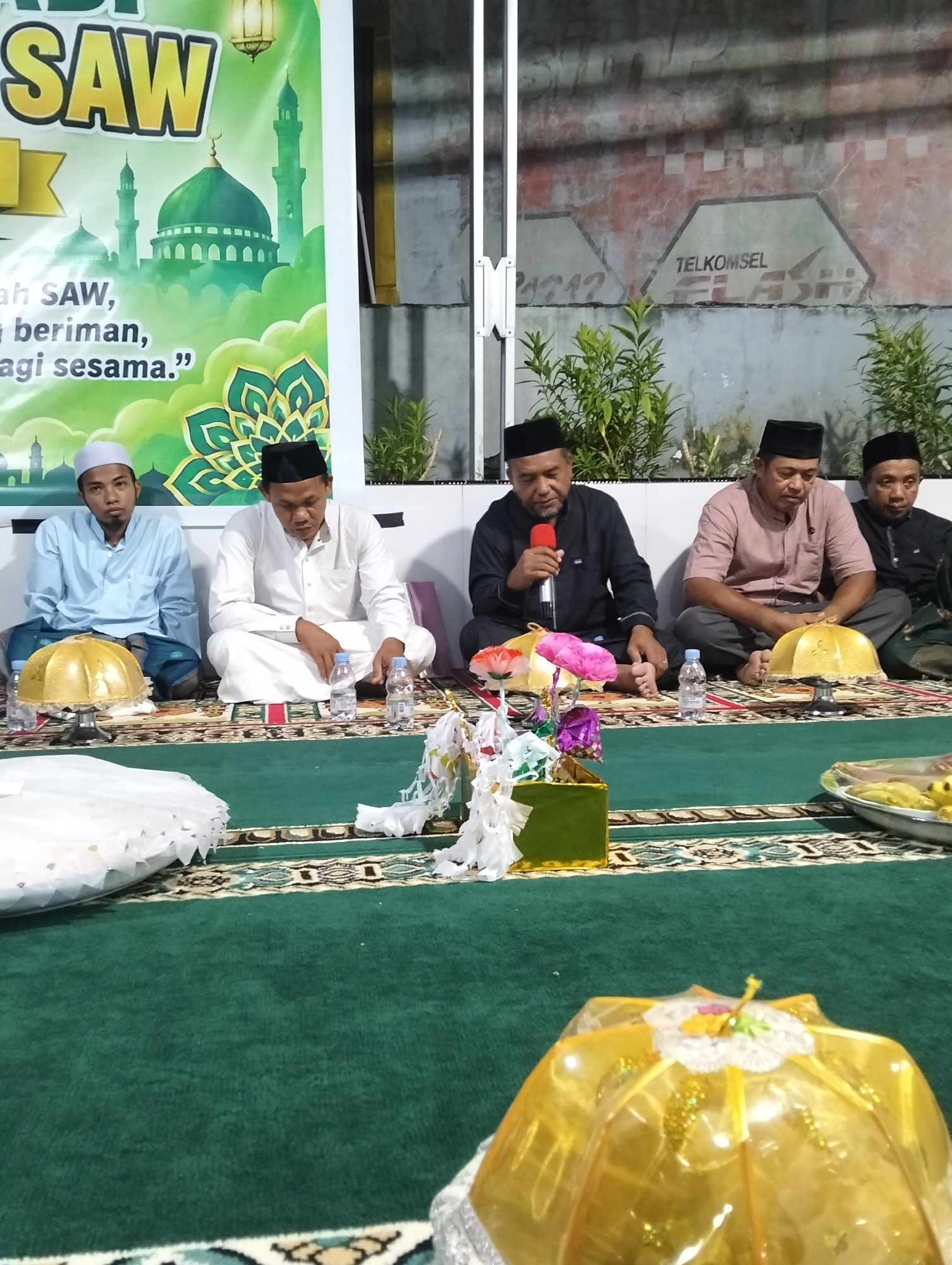 In Petapa, Indonesia, a group of men are seated on a green carpet indoors. Five men are visible in the foreground, all wearing traditional Islamic attire, including skullcaps or turbans. One man on the far left wears a light blue shirt and a white skullcap, while the man next to him is in a white garment and a black skullcap. The man in the center is holding a microphone and speaking into it, dressed in a black shirt and black skullcap. To his right, a man in a light brown shirt and black skullcap is seated, followed by another man in a dark shirt and black skullcap. Two bottles of water are placed in front of the men. Decorative items, including golden domes, colorful flowers, and white streamers, are arranged on the carpet. In the background, a banner with text and images of mosques is displayed. A wall with painted logos, including "TELKOMSEL FLASH," is also visible. Plants are seen growing in the background. The lighting suggests an indoor setting, possibly a community hall or mosque.