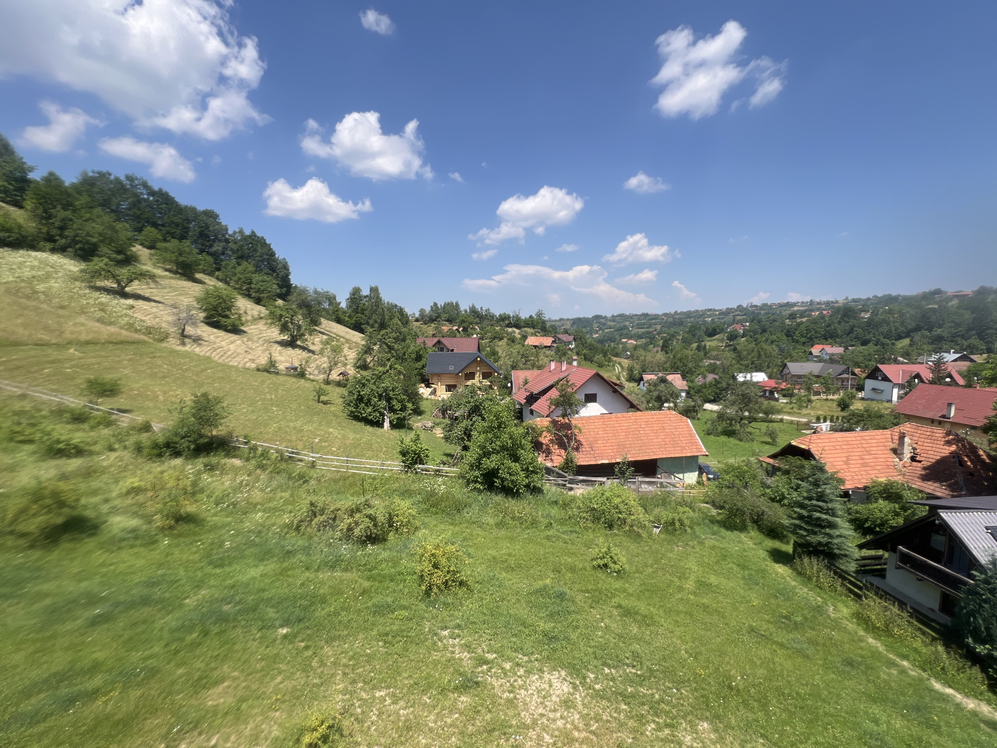 This is a wide-angle, eye-level outdoor shot of a rural village nestled within rolling green hills under a bright blue sky with scattered white clouds. The foreground is dominated by a lush, vibrant green meadow with patches of dry grass and small wildflowers. A light-colored wooden fence runs horizontally across the mid-ground, separating the meadow from a more cultivated area. Beyond the fence, several houses with terracotta tile roofs are visible, surrounded by trees and vegetation. The houses vary in size and architectural style, with some appearing to be traditional wooden structures and others more modern constructions. The village extends into the background, with more houses and trees scattered across the undulating landscape, leading to a distant treeline under the expansive sky. The overall impression is one of a tranquil, sun-drenched countryside scene.