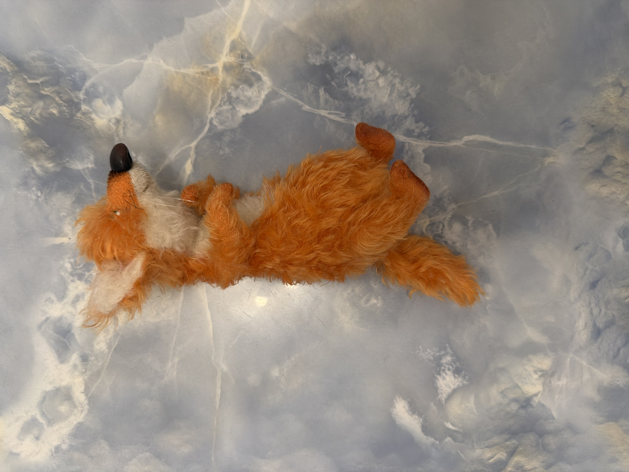 A stuffed fox toy lies on its back against a blue and white marbled background. The fox is orange with white fur on its belly, chest, and inner ears. Its nose is black, and it has thin whiskers. Its tail is bushy and orange. The background appears to be a smooth, possibly stone surface with irregular veining. The lighting is soft and diffused, casting a slight shadow beneath the toy.